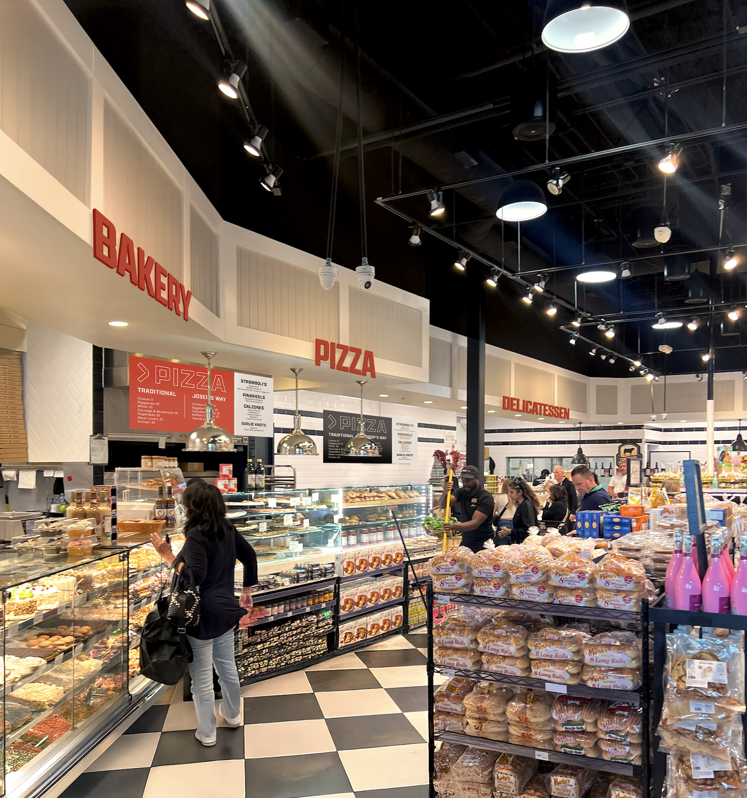 Interior of a bakery and deli with customers shopping for baked goods, deli meats, and bread, featuring signs for bakery, pizza, and delicatessen sections