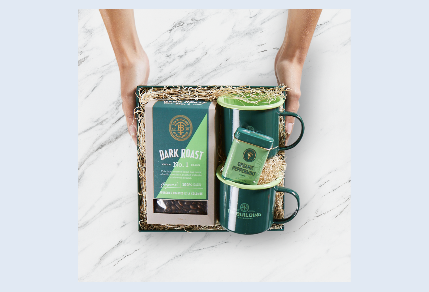 A gift box with a box of dark roast coffee, two green mugs, and a mint flavored organic peppermint tea tin, set on a white marble surface. Robert Ranfranz branding, design and ecommerce.