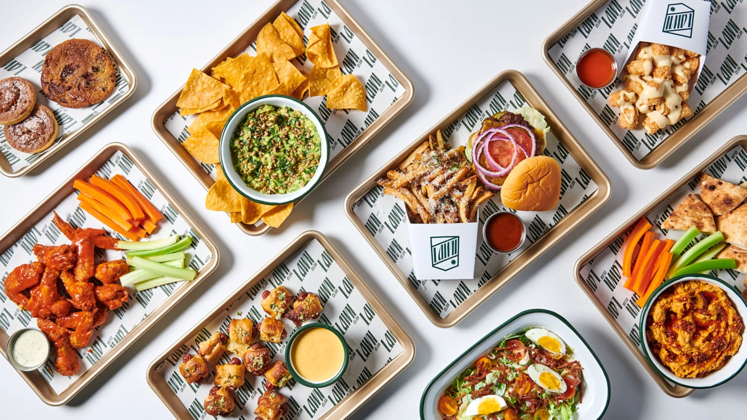 Multiple trays of assorted Mexican dishes including nachos, tacos, chicken wings, dips, fried items, and salad, arranged on a white surface.