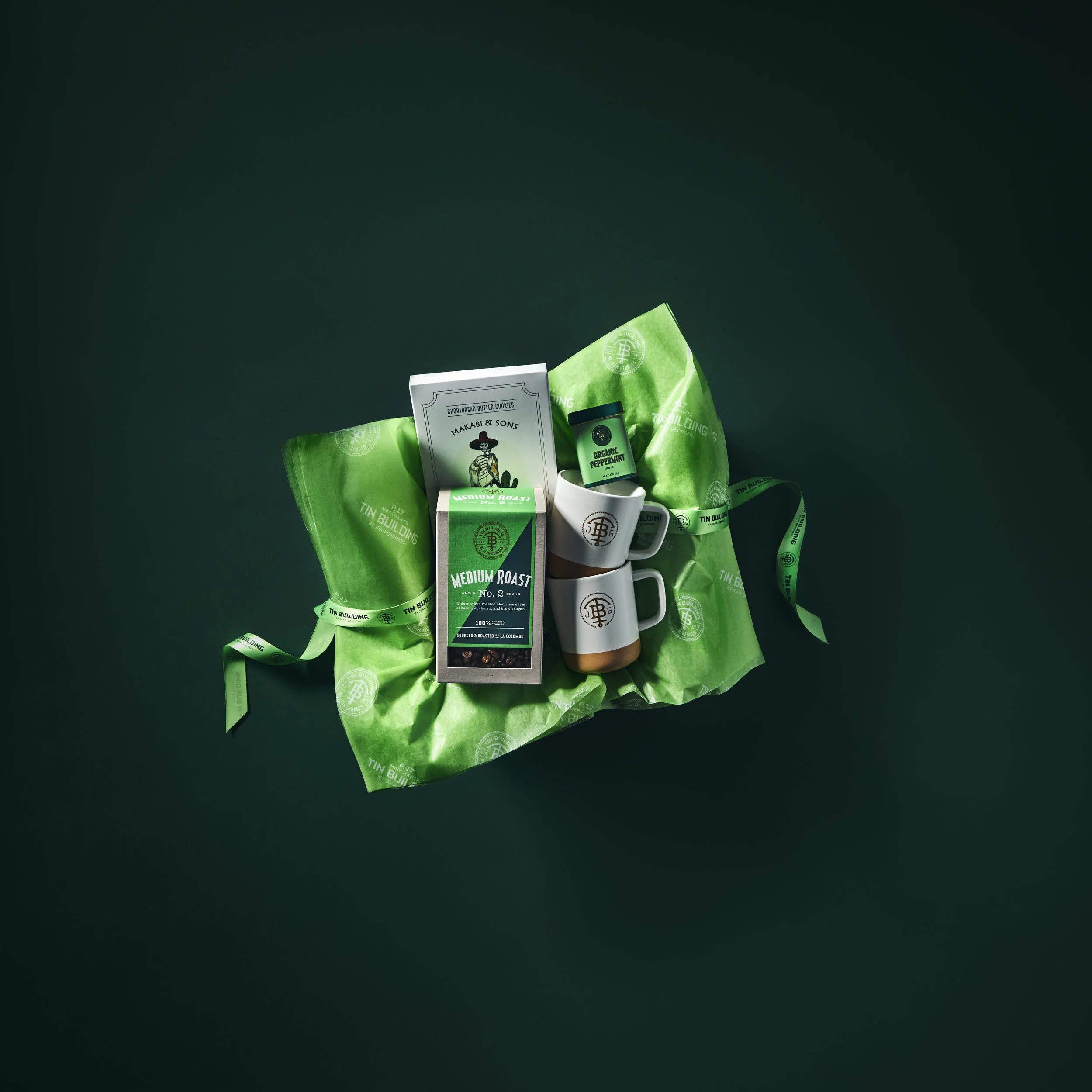 Various green and white packaged products, a white mug, and a small green container arranged on a dark surface, with a green ribbon and a blank string tag nearby. Robert Ranfranz branding, design and ecommerce.