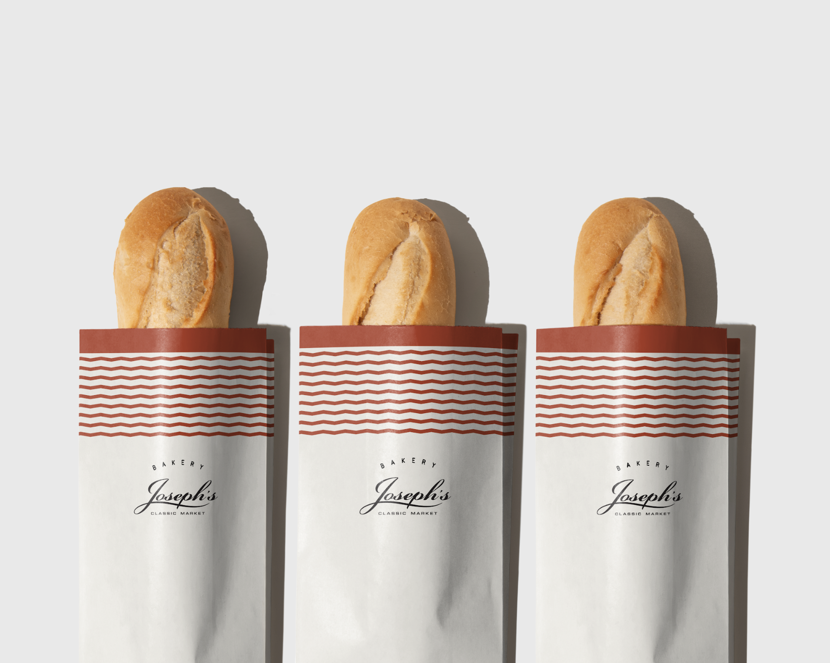 Three baguettes in white paper wrappers with red and white wavy stripe design, labeled 'Bakery Joseph's Classic Market,' against a plain light background.
