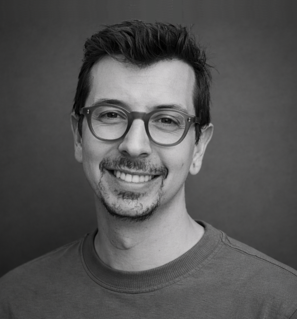 A black and white portrait of a man with glasses smiling at the camera, wearing a crew neck shirt, with a dark plain background.