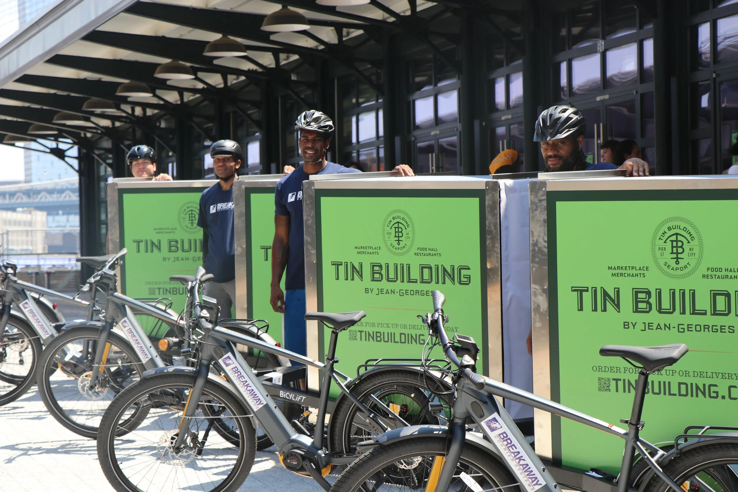 Four people wearing helmets and blue shirts standing behind green carts with bicycles parked in front. The background shows a glass building with an overhang and a bridge in an urban setting.