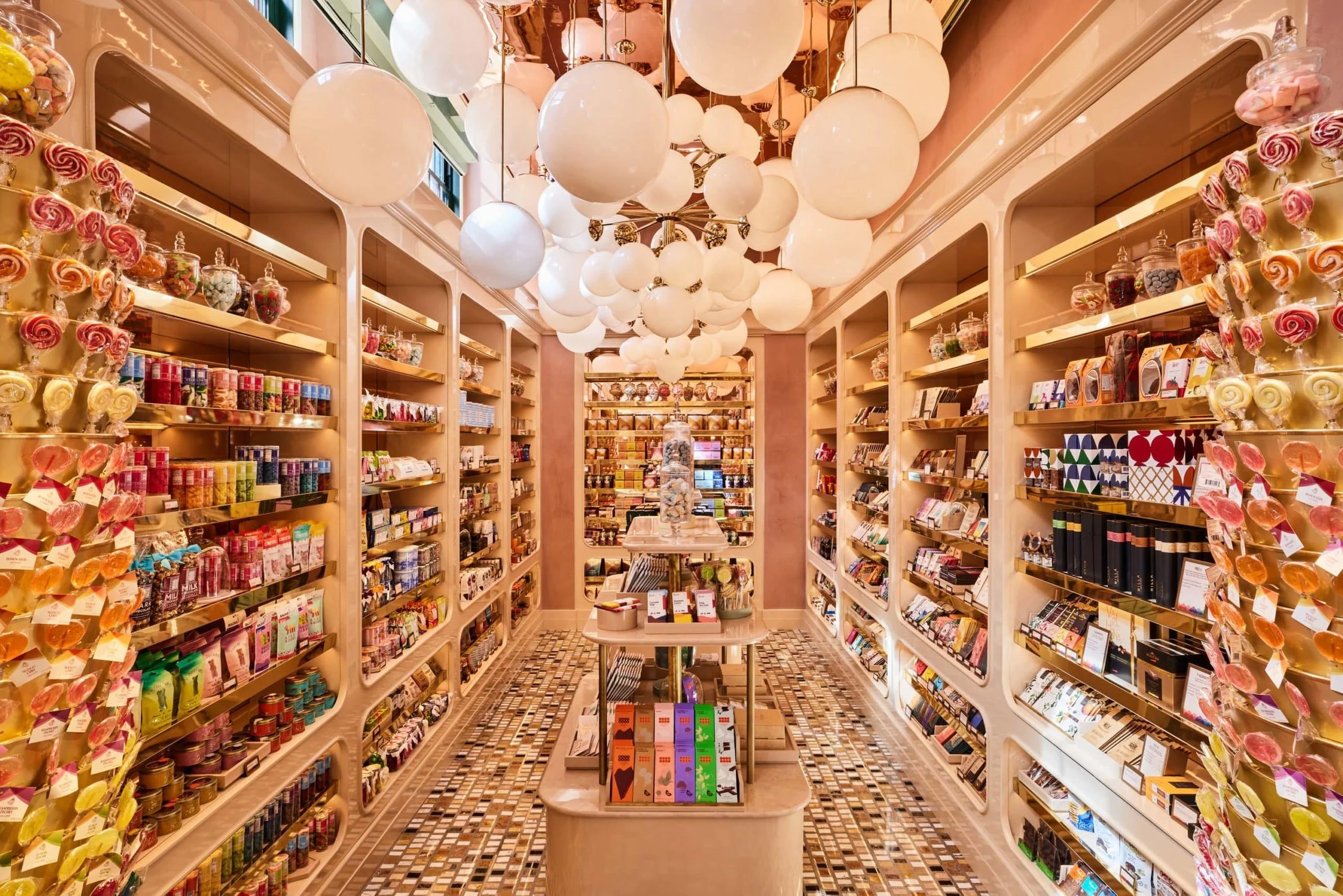 A candy store aisle with shelves filled with colorful candies and sweets, illuminated by large white globe pendant lights hanging from the ceiling.