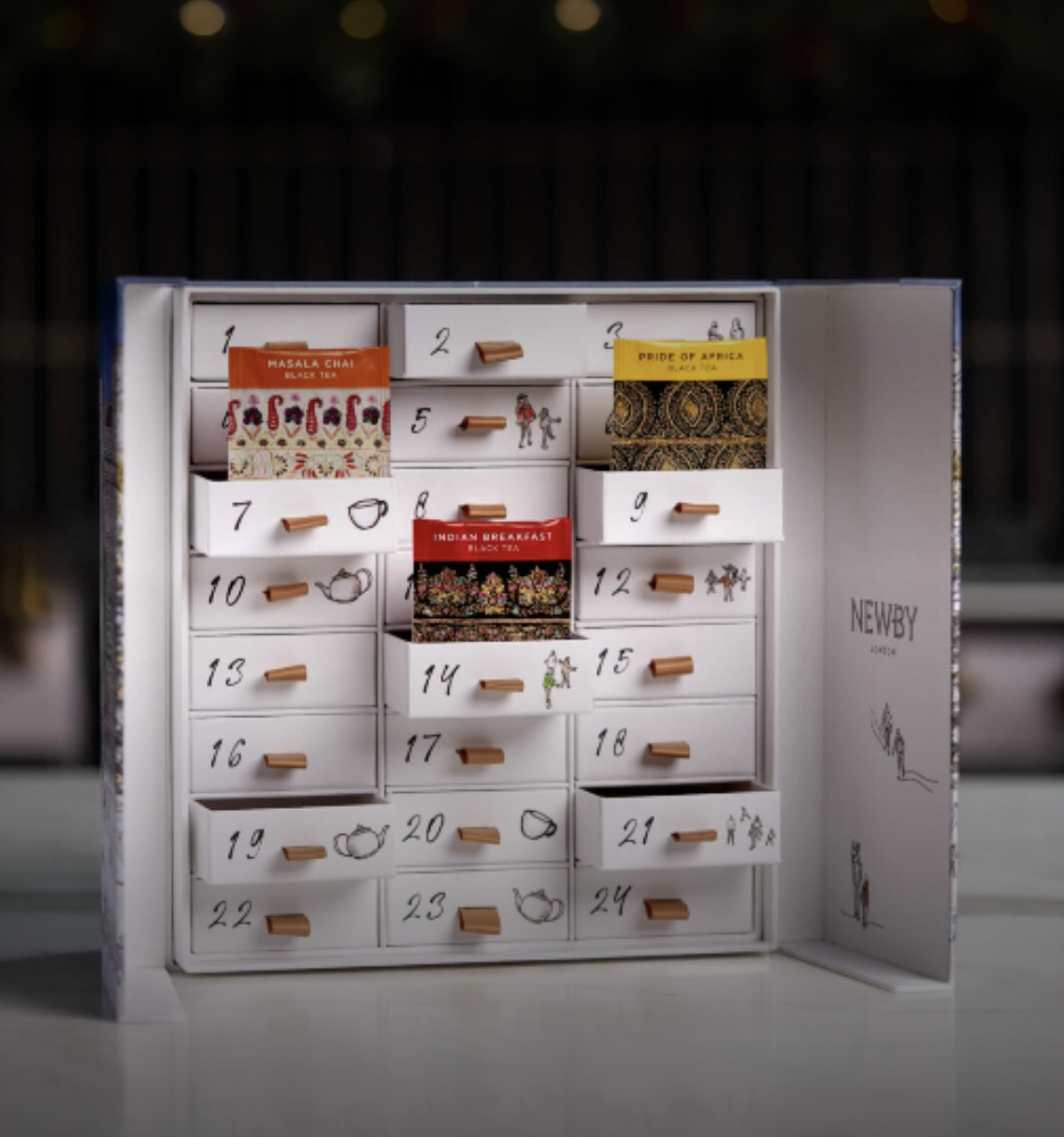 An ornate advent calendar with small numbered drawers, each decorated with illustrations of tea cups and people, containing various tea packets from different brands such as Masala Chai, Pride of Africa, and Indian Breakfast.