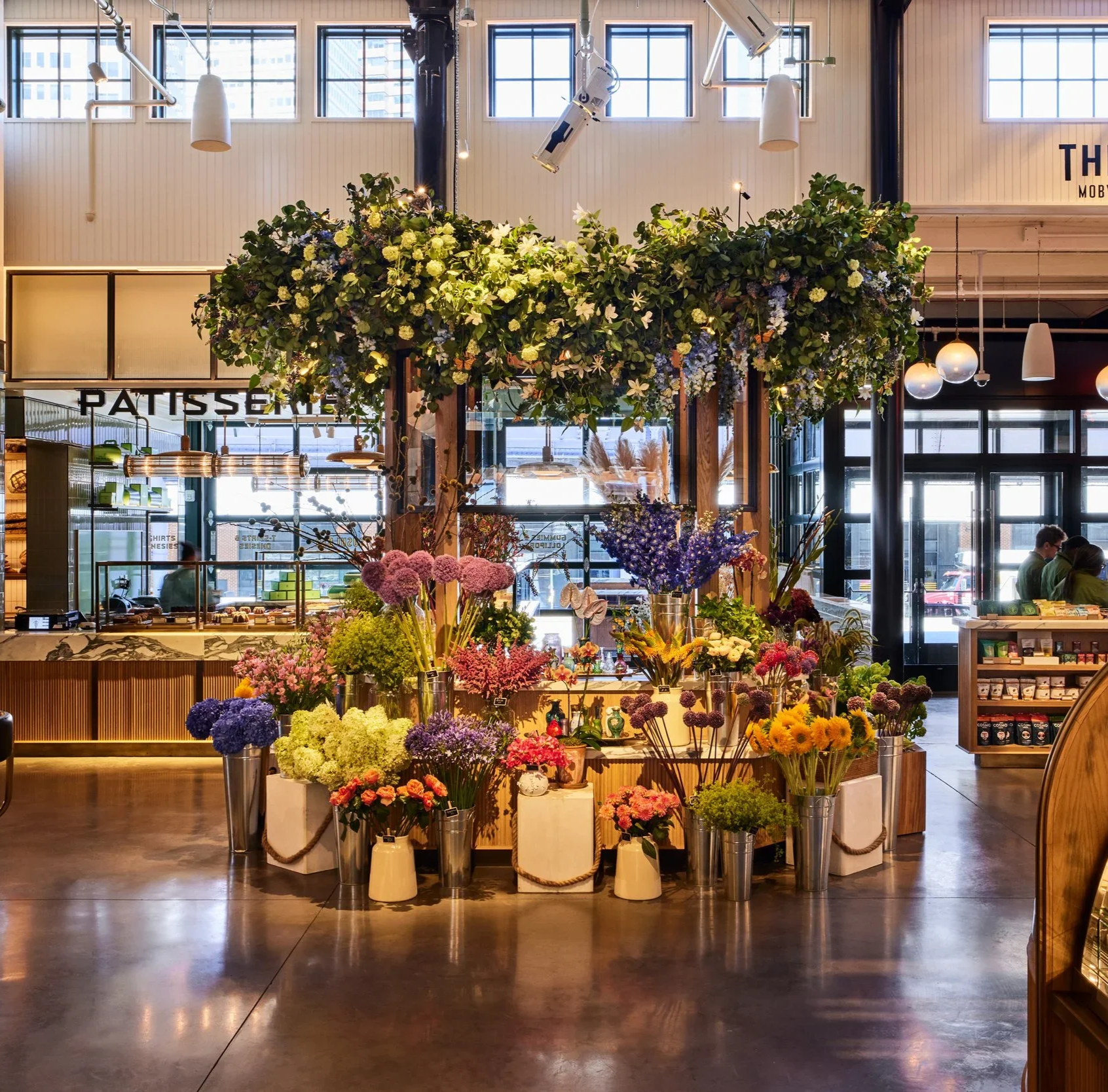 A flower display with various colorful flowers in vases inside a modern indoor space, with a wooden framing structure and hanging greenery overhead.