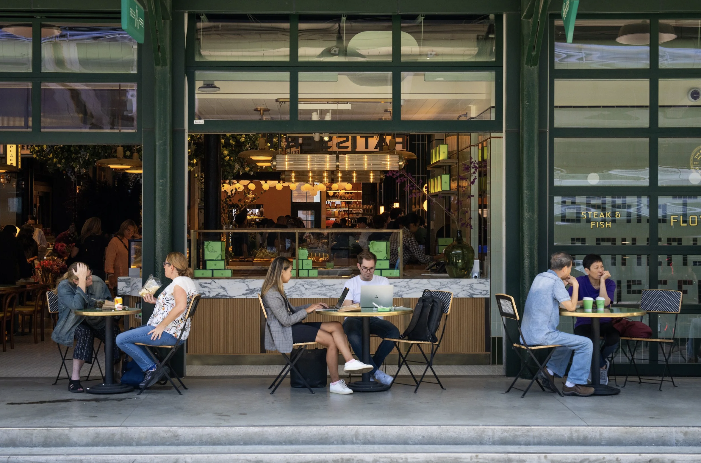 People sitting at tables outside a restaurant with a modern design, large windows, and green framing, dining and using laptops. Robert Ranfranz branding, design and retail.