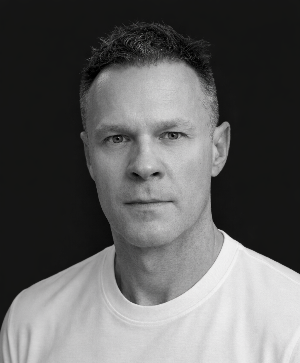 Black and white portrait of a man with short, textured hair, wearing a white t-shirt, looking directly at the camera against a dark background.