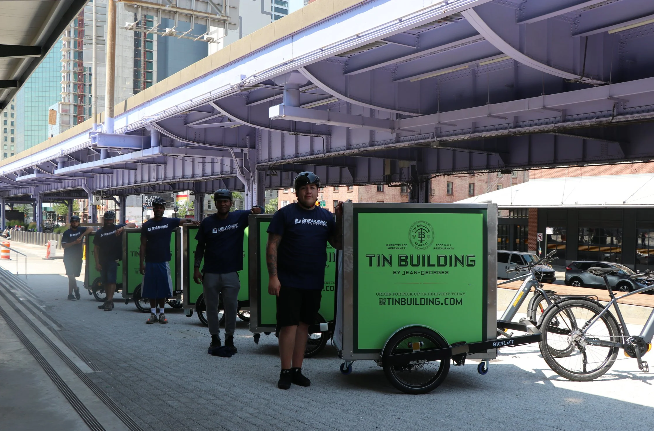 A group of delivery workers standing next to green delivery carts with bicycles attached, on a city sidewalk under an elevated train track.