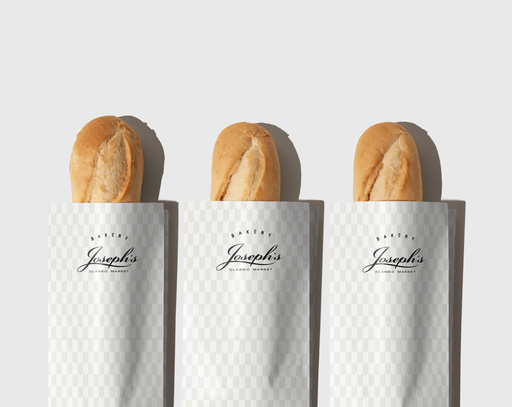 Three bakery breadsticks in white wrapping paper with checkered pattern, labeled 'Joseph's Bakery Classic Market,' against a plain light grey background.