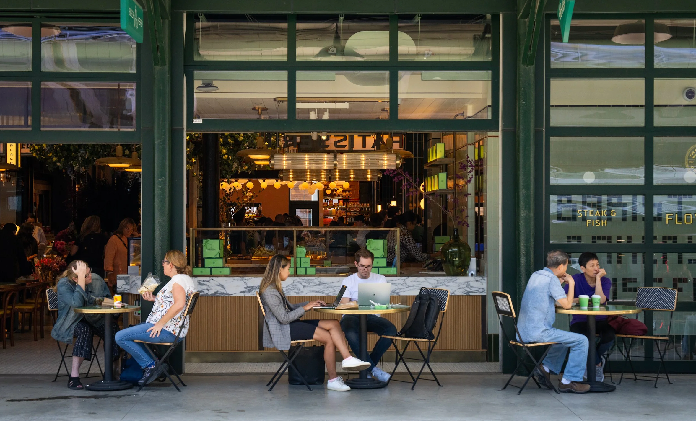 People dining and working outside at a restaurant with large glass windows and a colorful interior visible in the background. Robert Ranfranz retail services.