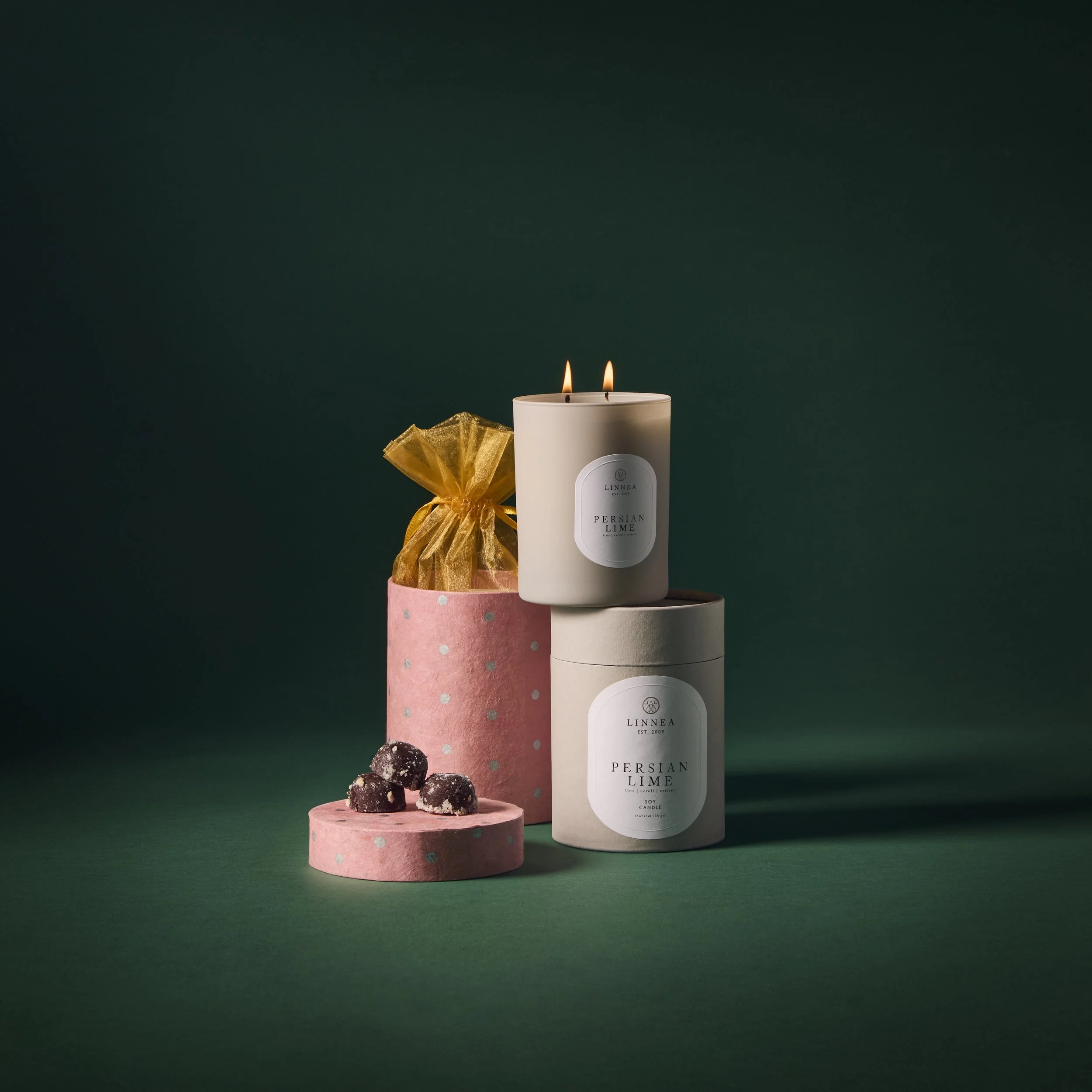 Collection of scented candles, gift box with chocolates, and decorative storage boxes on dark green background.