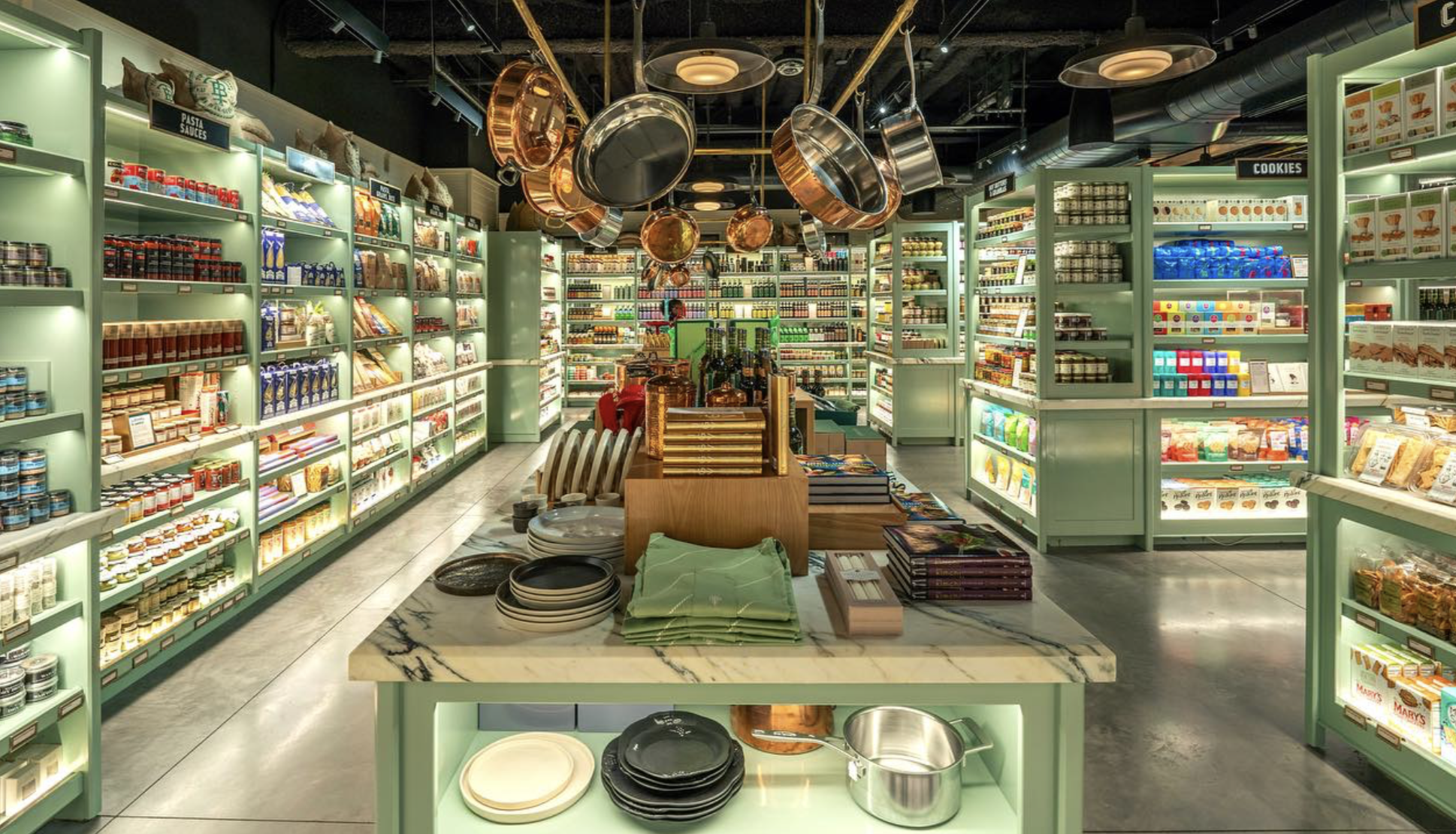 Interior of a retail store with green shelving units stocked with various food products, and a central display table with plates, bowls, and kitchenware, under hanging copper pots. Robert Ranfranz branding, design and retail.