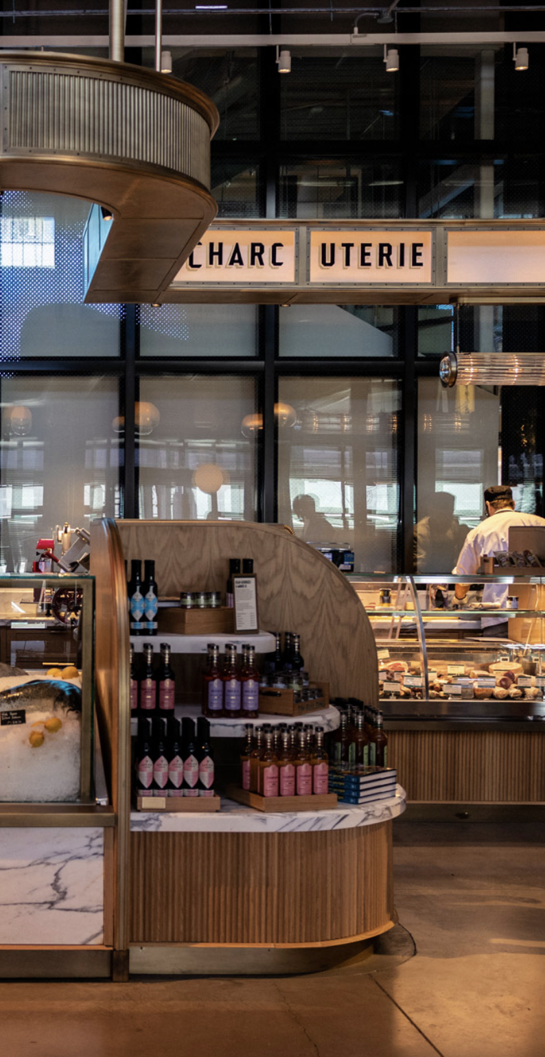 Interior of a modern restaurant or cafe with display cases of food and beverages, visible staff working behind the counter, and a sign with partially obscured text.