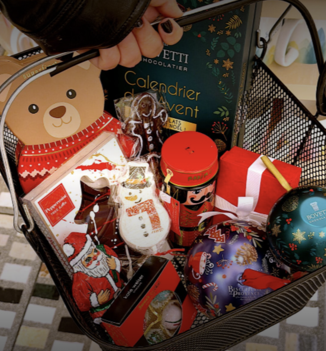 A shopping basket filled with Christmas-themed items, including a teddy bear, a gingerbread man, Santa and snowman cookies, decorative ornaments, and a Christmas calendar.