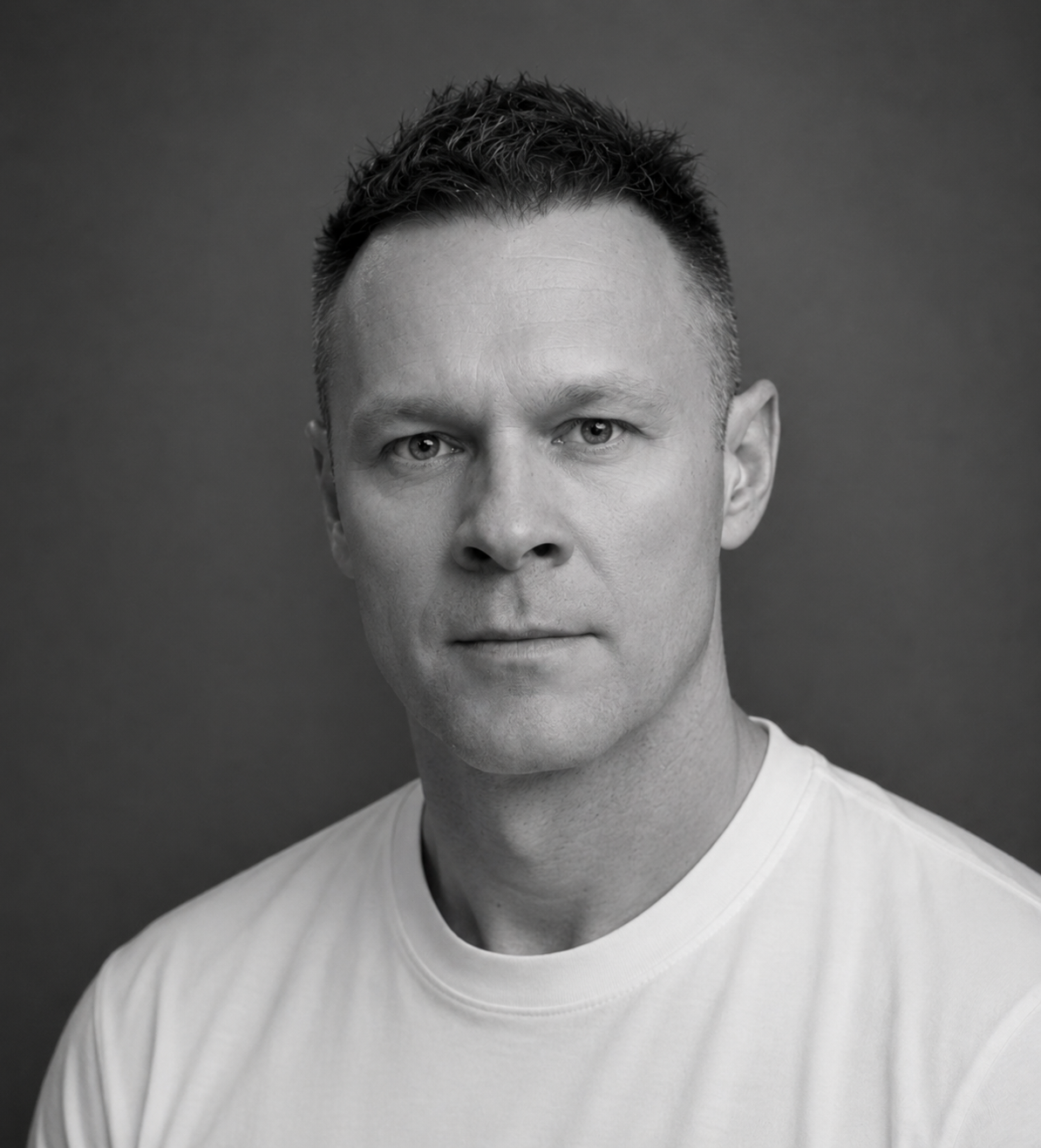 Black and white portrait of a man with short, curly hair, wearing a white t-shirt, against a dark background.