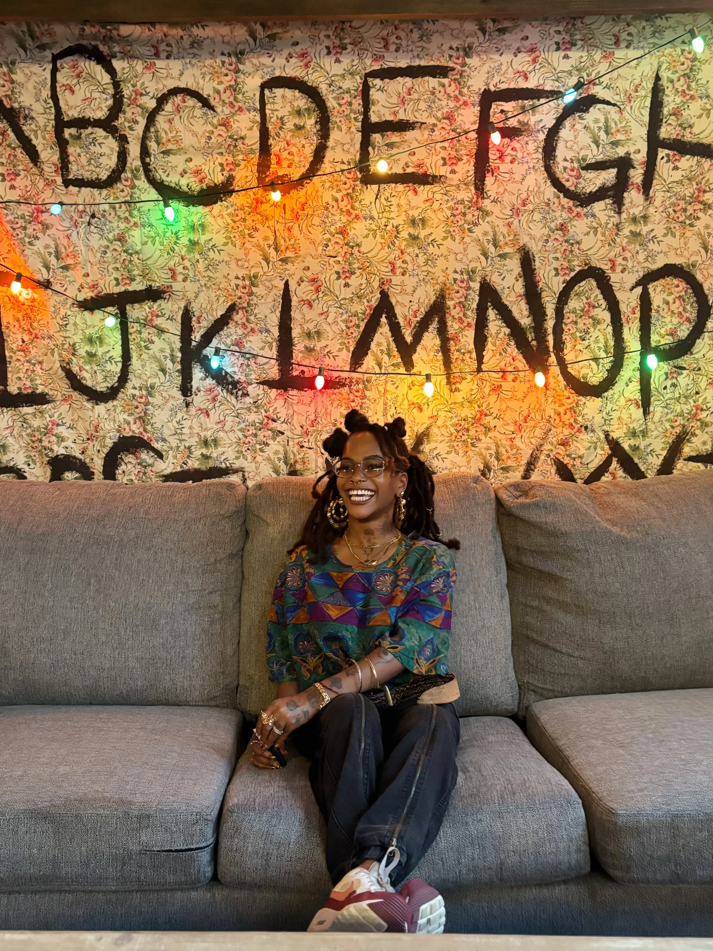 A smiling person sitting on a gray couch with legs crossed, wearing glasses, a colorful patterned shirt, and multiple gold jewelry pieces, in front of a floral-patterned wall with multicolored string lights.