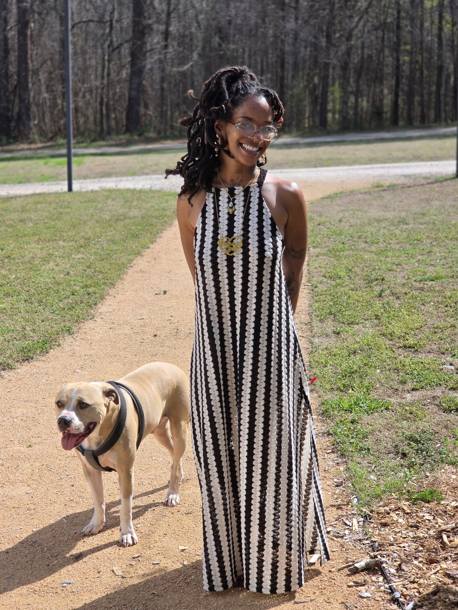 A woman with dreadlocks wearing glasses and a black and white striped dress stands outdoors on a dirt path with a dog on a leash in front of her. The background shows trees and greenery.