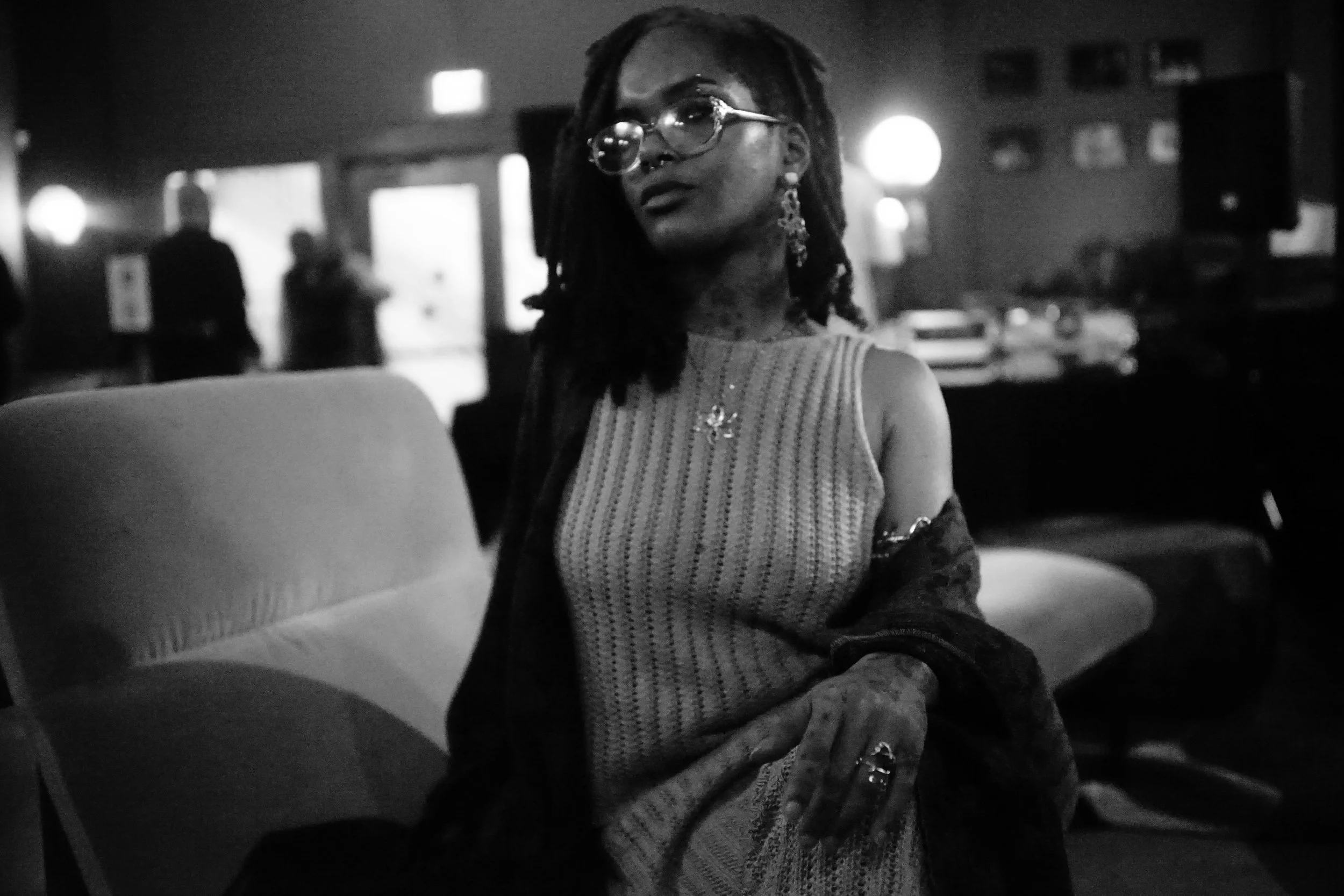A woman with dreadlocks and glasses sitting on a chair in a dimly lit room, wearing a striped sleeveless top and earrings, with blurred figures in the background.