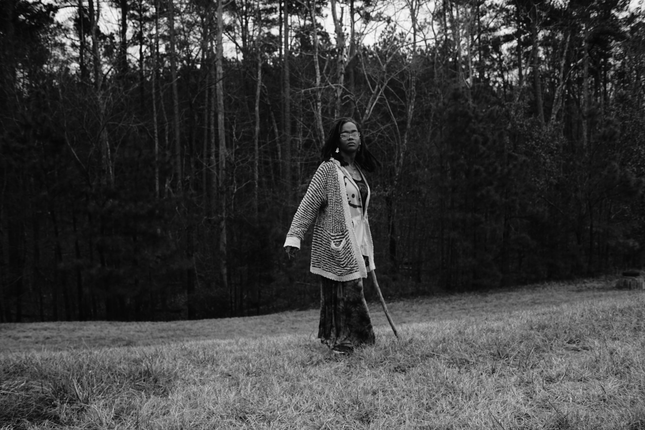 A woman wearing glasses and a patterned cardigan standing in a grassy area with trees in the background, holding a walking stick, in a black and white photograph.