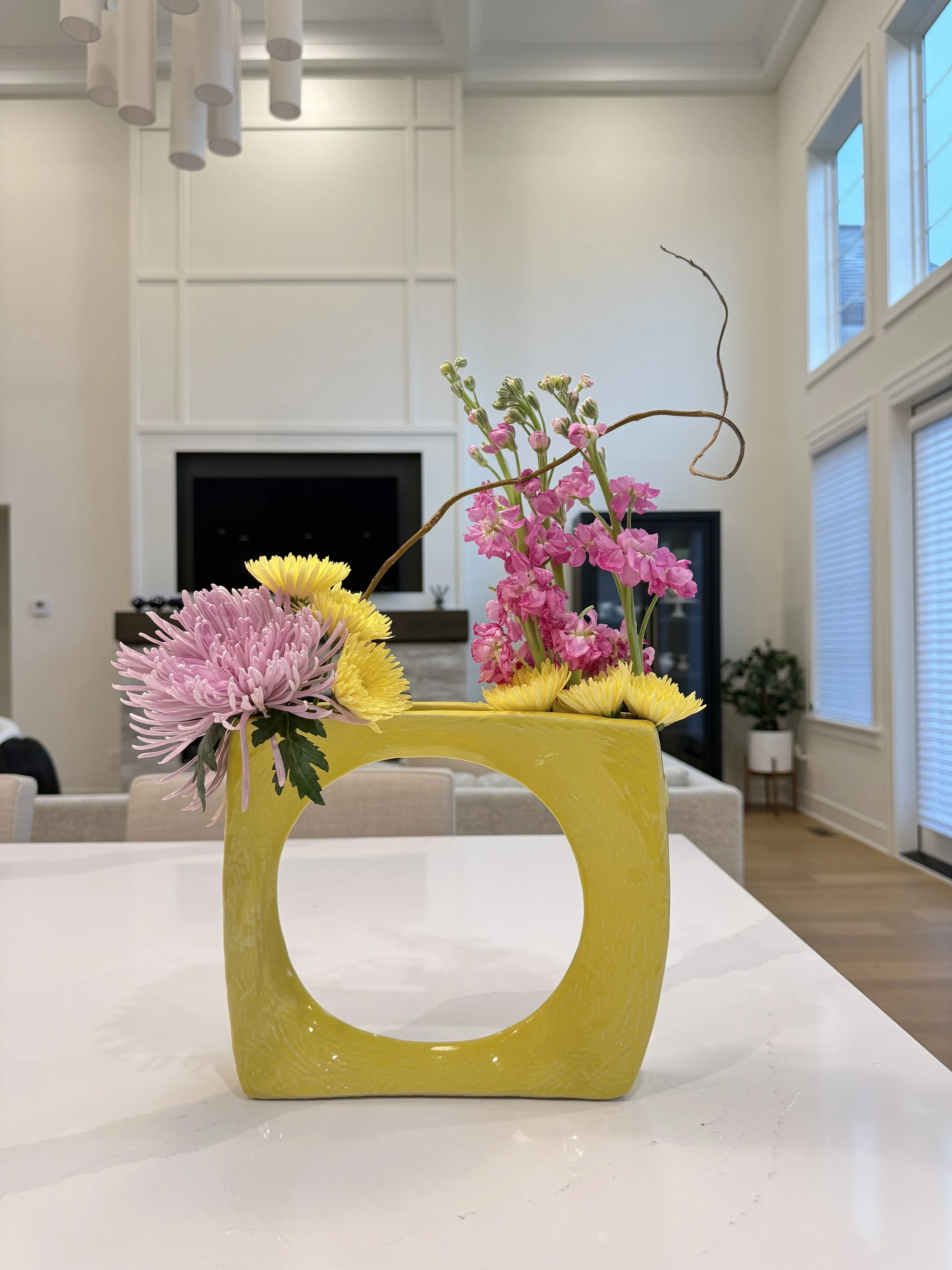 A yellow ceramic vase with a circular handle in the middle, holding pink, yellow, and purple flowers with green leaves, placed on a white table in a bright living room with large windows and a fireplace.