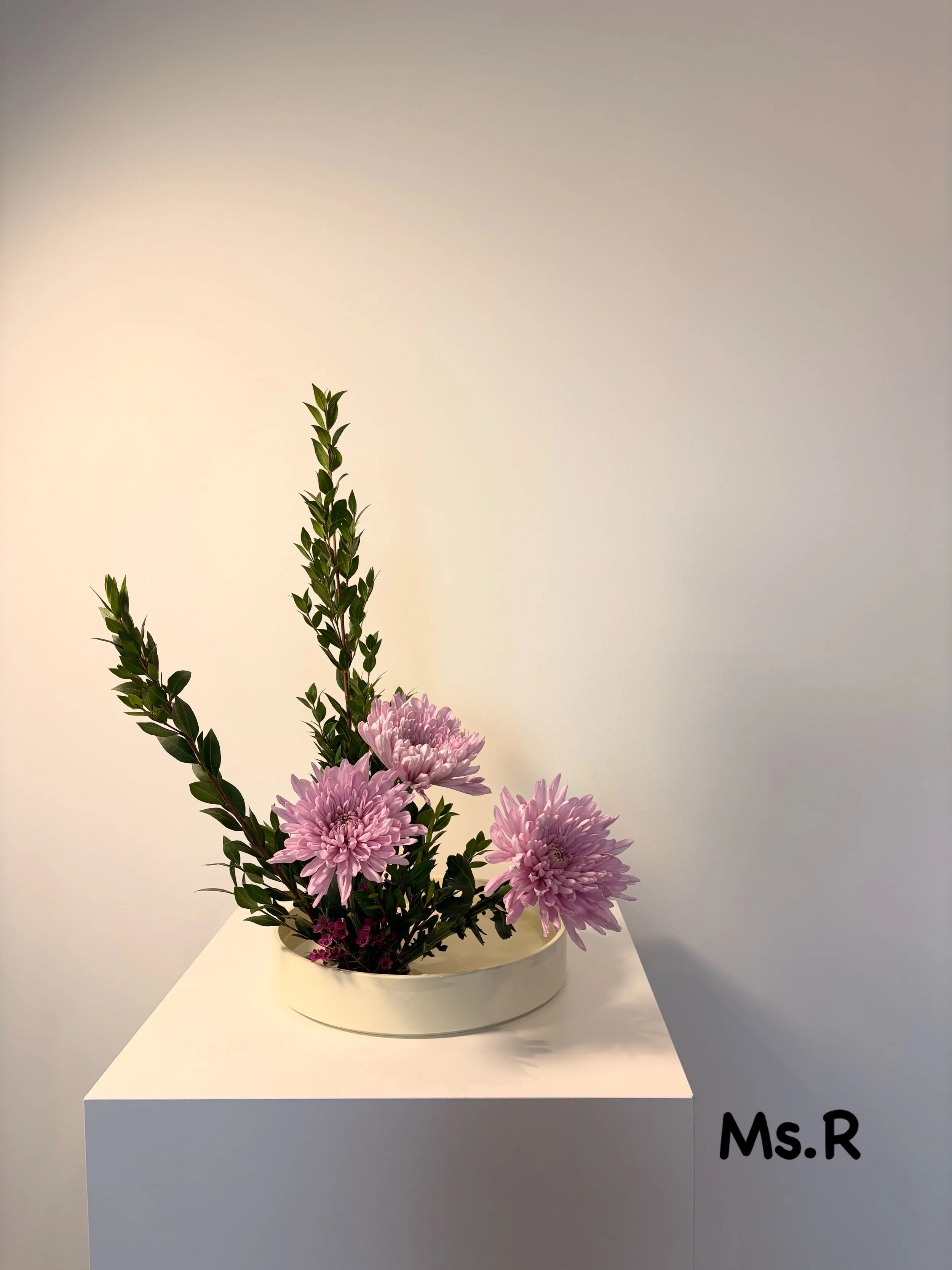 A floral arrangement with pink chrysanthemums and green foliage in a white oval vase on a white pedestal against a plain light-colored background.