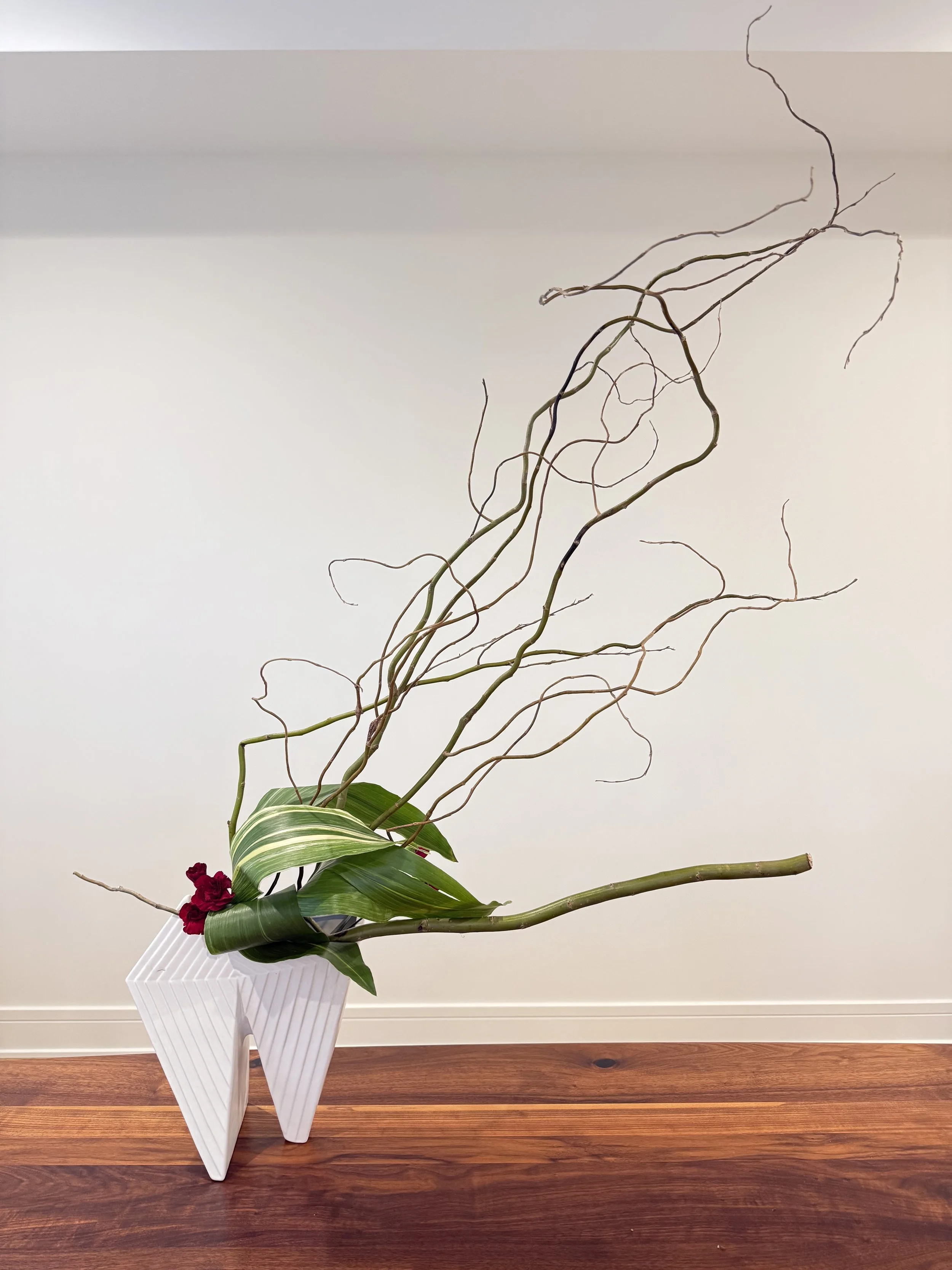 Decorative plant arrangement in a unique white geometric vase on a wooden floor, with a light-colored wall in the background.