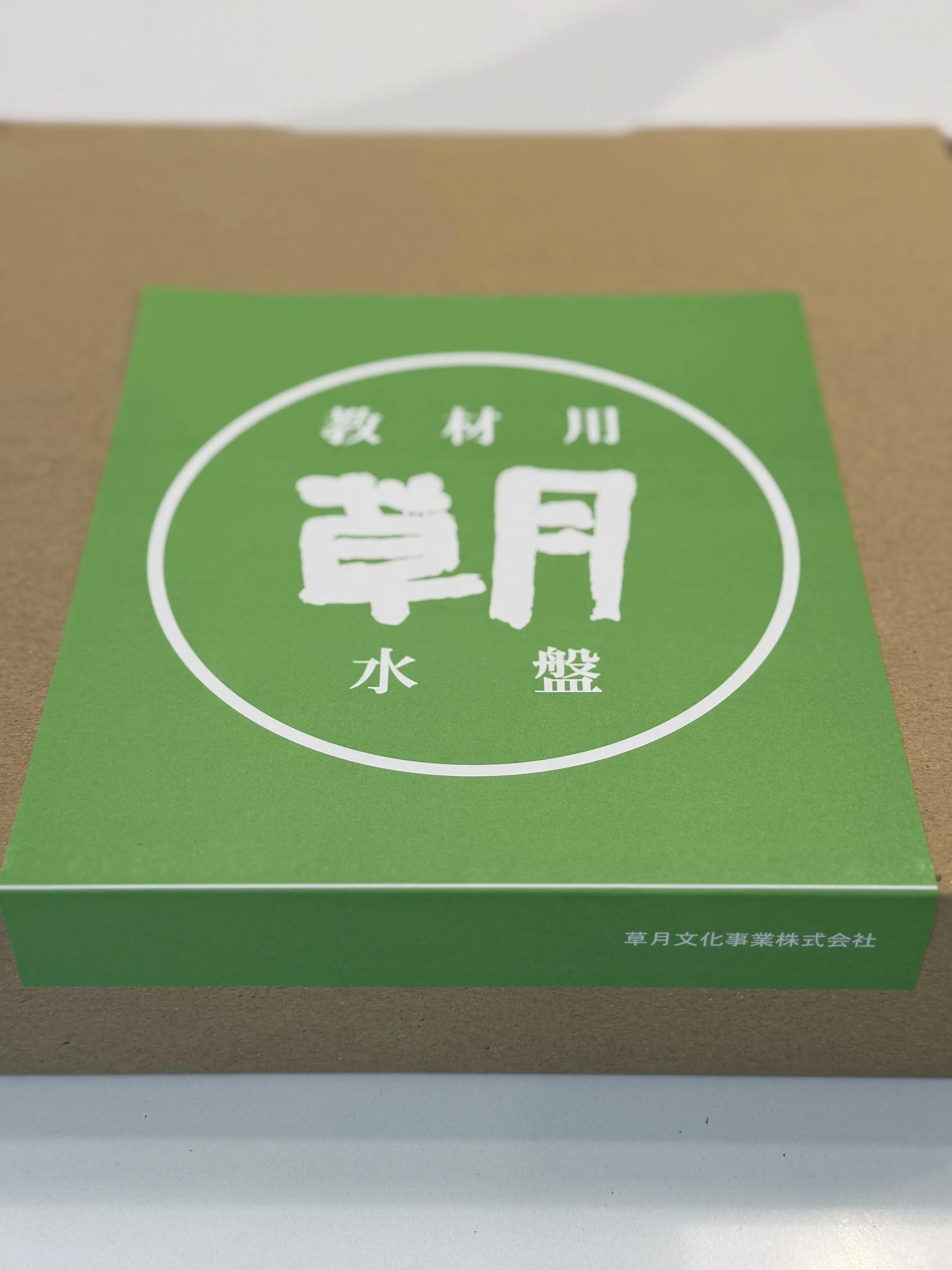 A green box with white Japanese text and a white circle design on the top. The box is placed on a beige surface, with additional Japanese text at the bottom.
