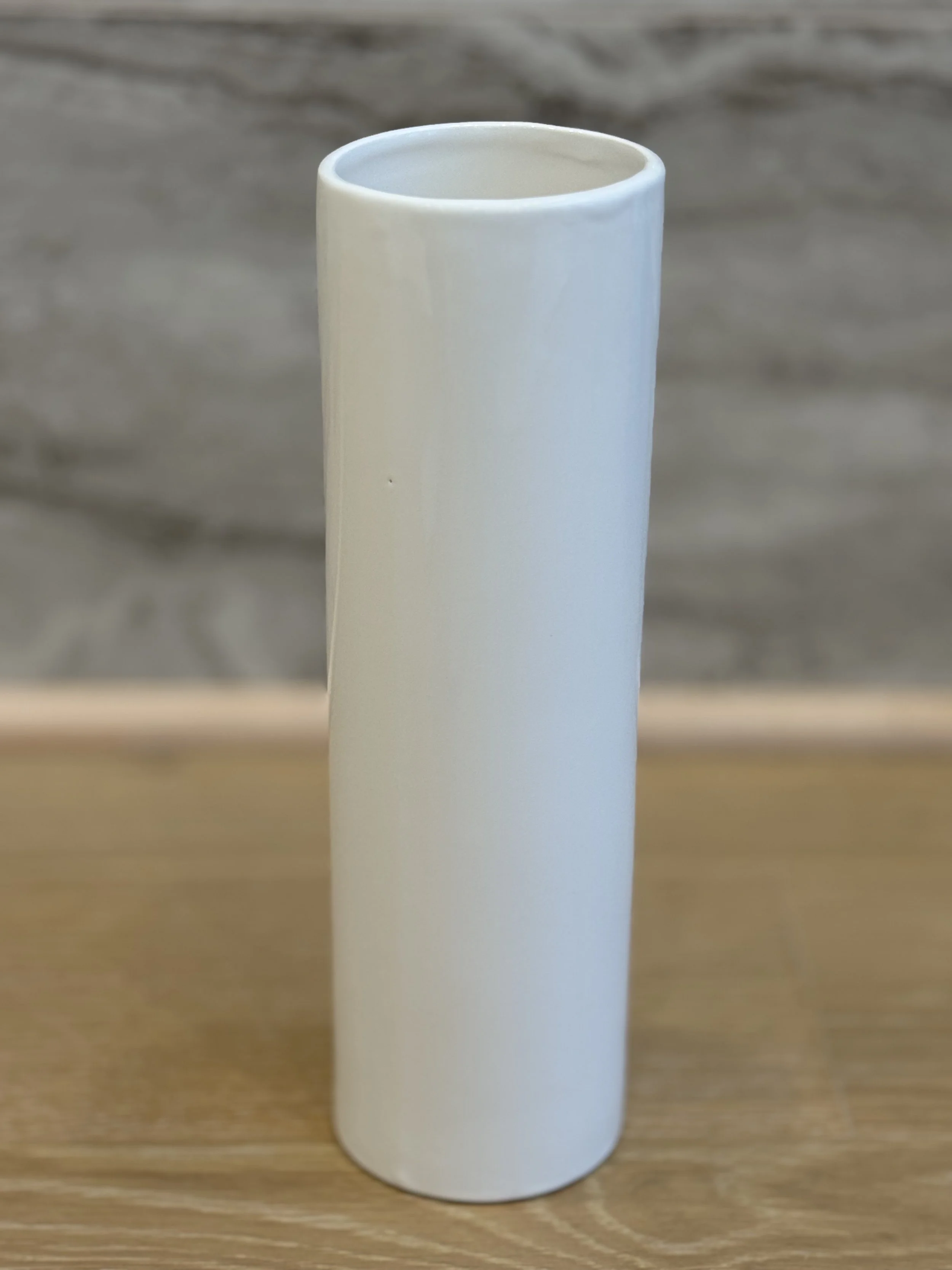 A plain white cylindrical ceramic mug with no handle, sitting on a wooden surface with a blurred wooden background.