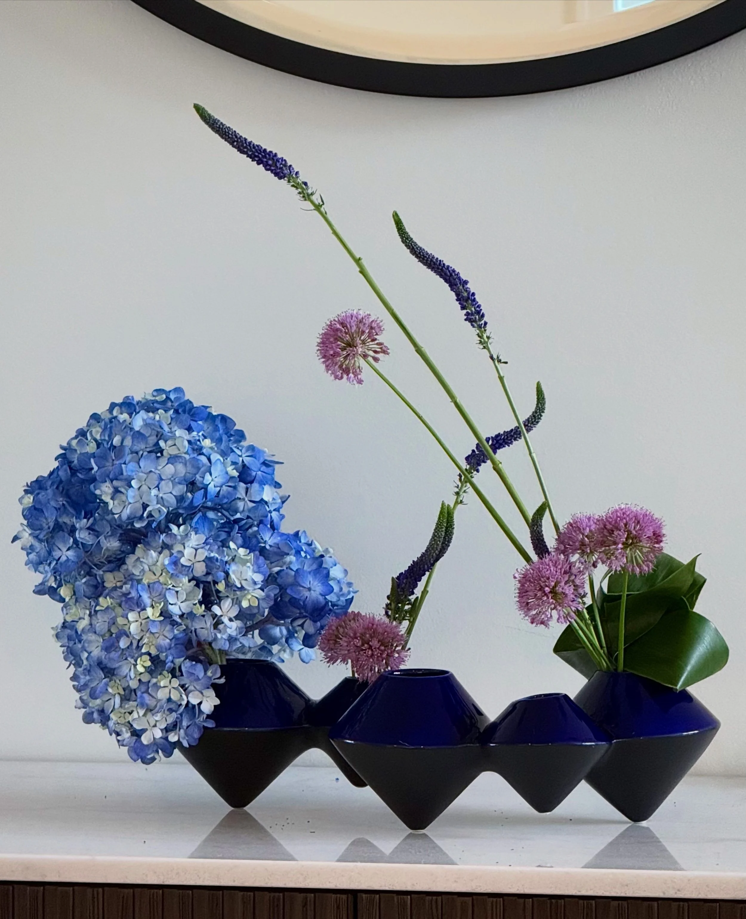 A black decorative vase with a geometric pattern holding a floral arrangement of blue hydrangeas, pink allium, and tall purple veronica flowers, placed on a white countertop in front of a white wall with a round mirror.