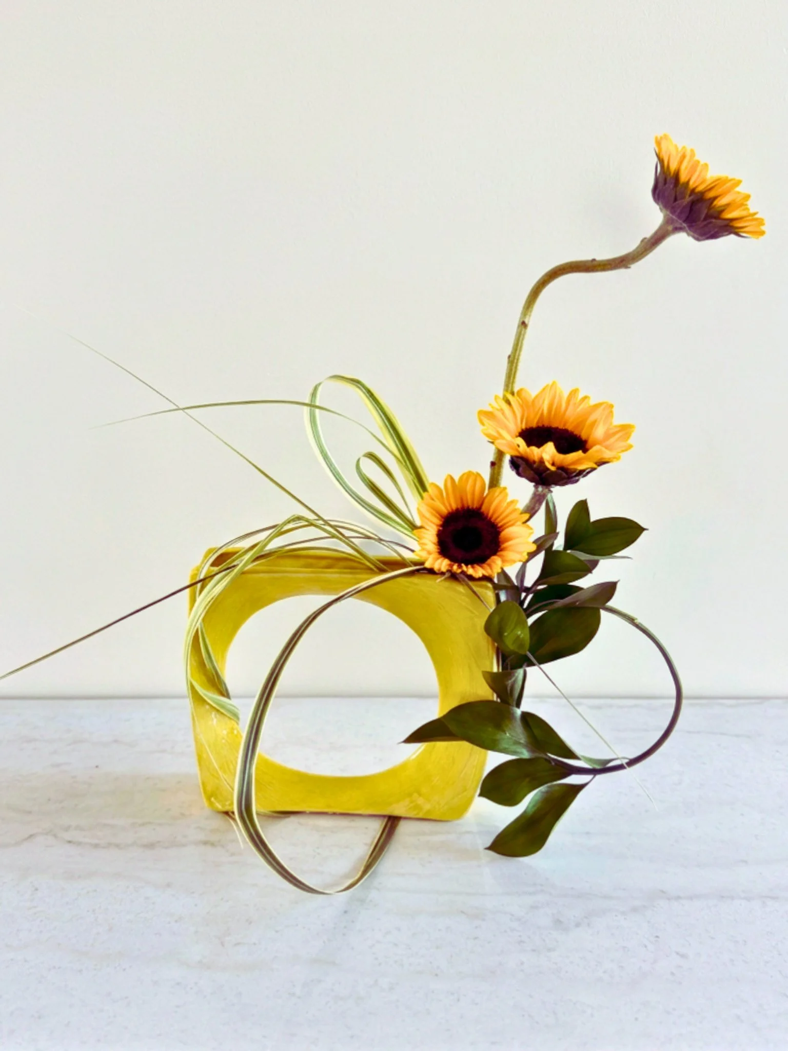 Yellow ceramic vase with orange and yellow sunflowers and green leaves, white background.