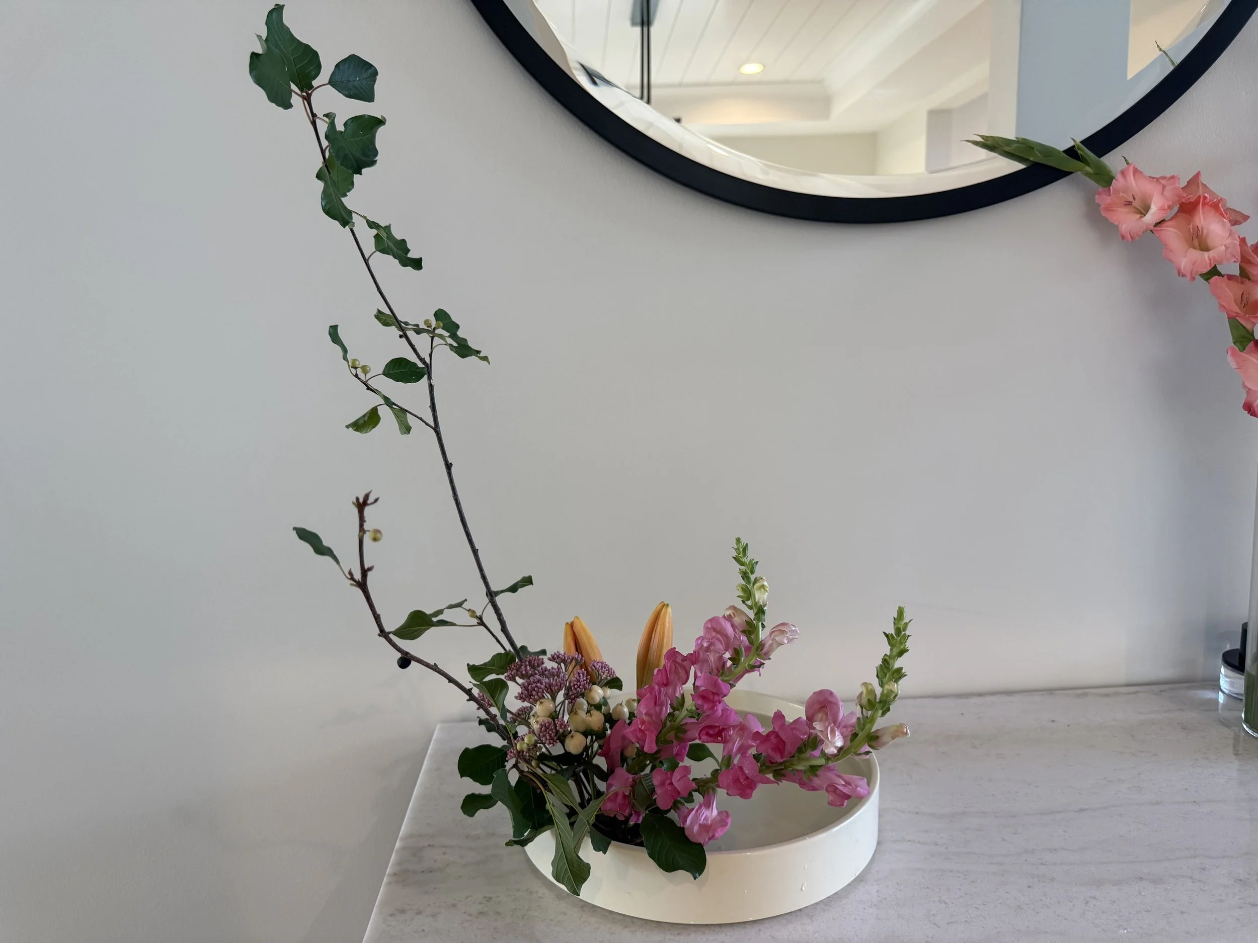 A flower arrangement in a white oval vase on a light-colored countertop. The arrangement includes pink gladiolus, pink snapdragons, greenery, and small white berries. A round mirror is on the wall above the arrangement, reflecting a ceiling and part of a room.