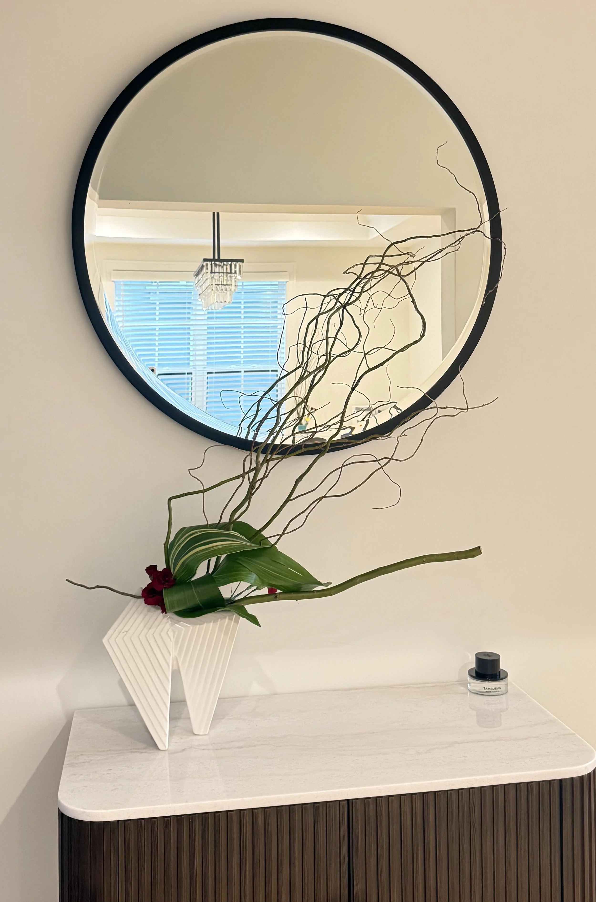 A round wall mirror reflecting a room with window blinds and a chandelier. In front of the mirror, there is a white geometric vase with a green plant and a red flower. The surface below the vase has a small jar labeled 'TAMBURINS' and a black container.