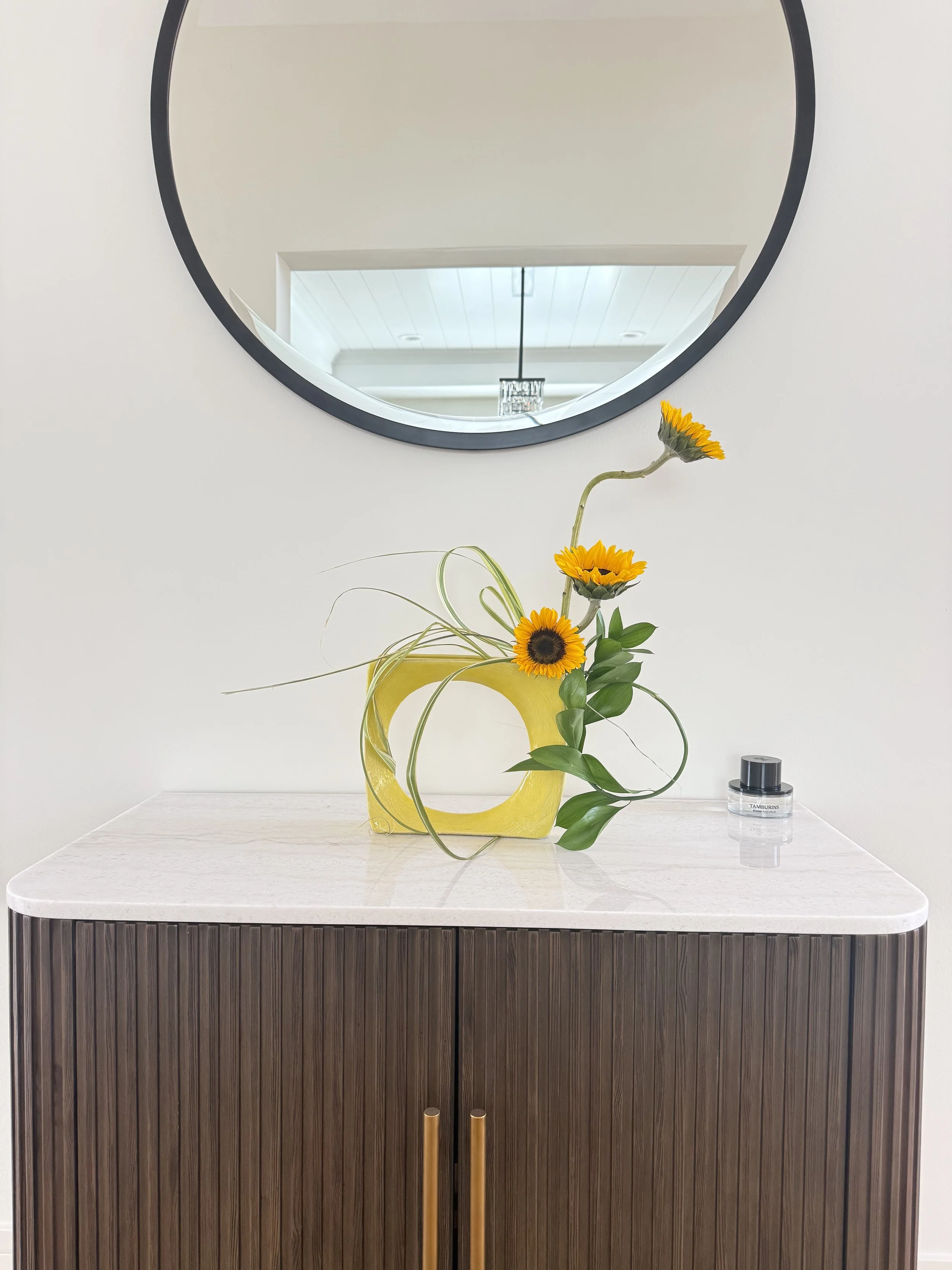 A yellow vase with a curved handle holding sunflowers and green foliage is placed on a white countertop. A large circular mirror is mounted on the wall above, reflecting a ceiling with a chandelier and a white wall.