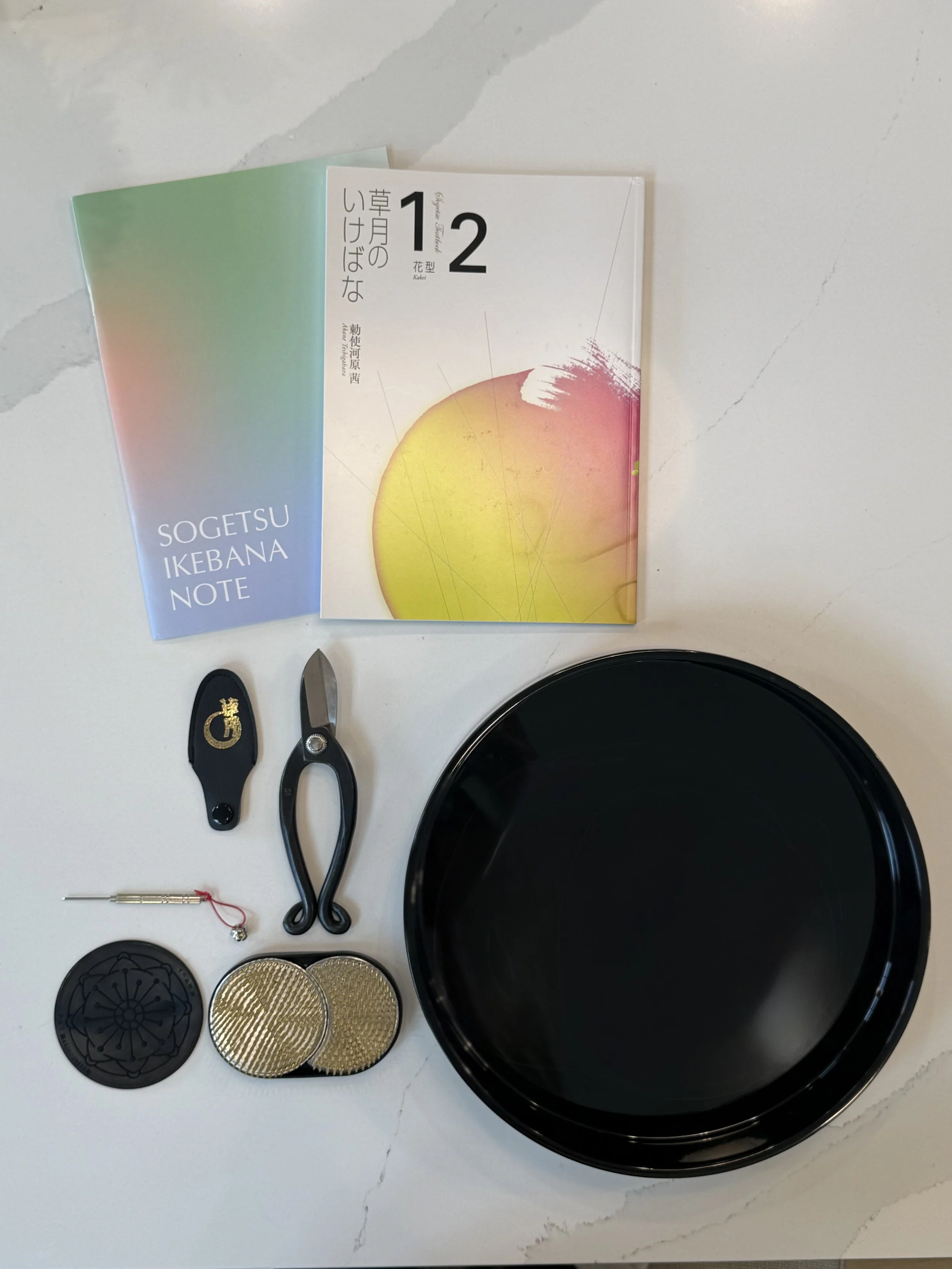 A collection of tools and items on a white marble surface, including a pair of black scissors, a small screwdriver, decorative coins, a black flower-shaped container lid, a gold coin, a black container lid, and a black round tray. There are also two booklets, one with a gradient cover and the other with a white floral design, and a pair of shoes with a gold emblem.