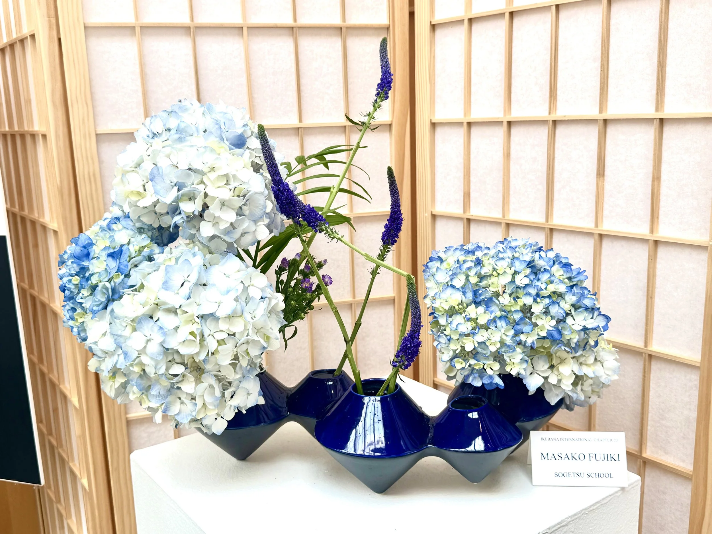 A floral arrangement in a blue ceramic vase with blue and white hydrangeas and tall purple flowers on a table. A wooden lattice backdrop and a white sign with black text are visible.