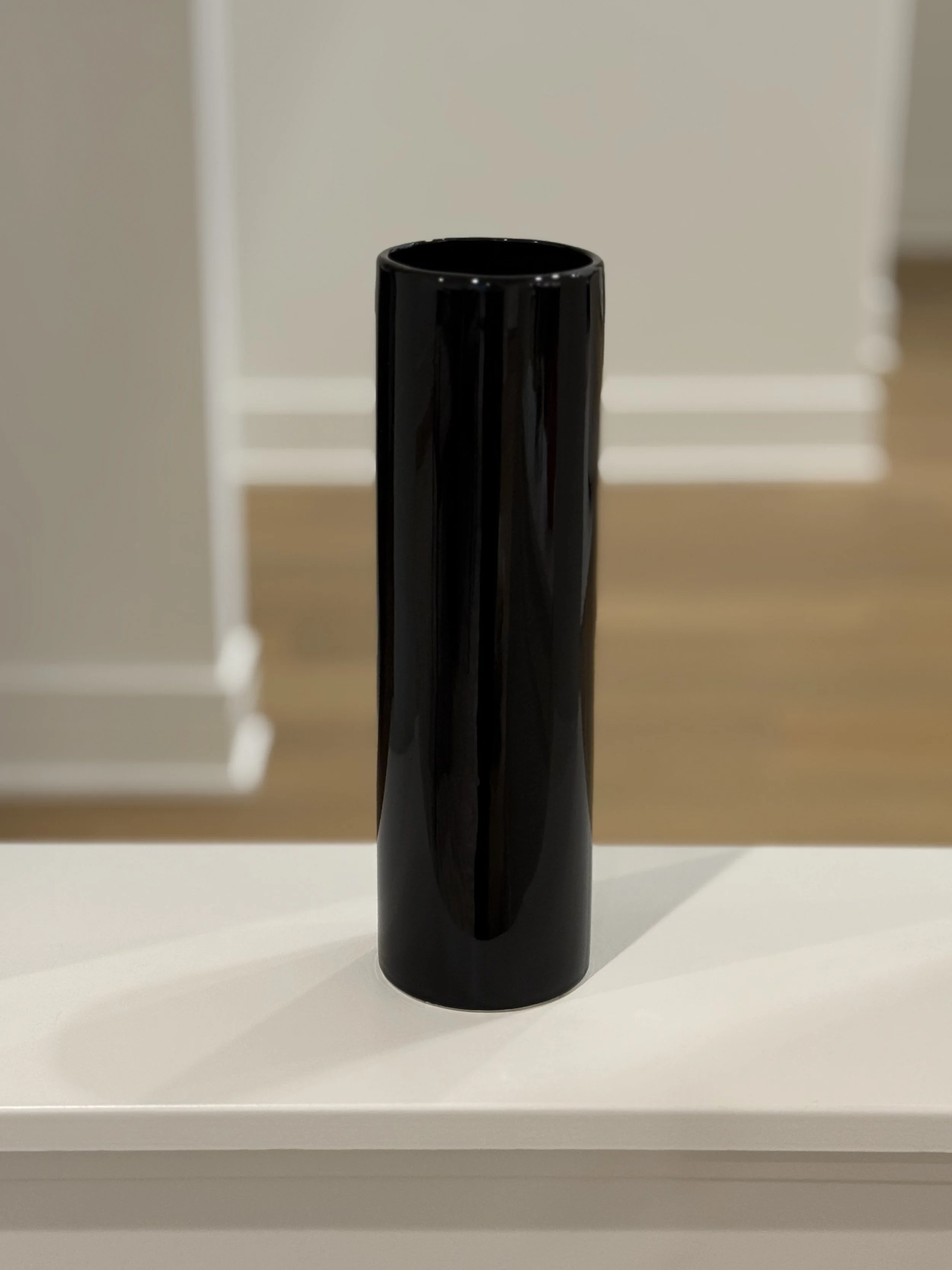 A black cylindrical object with a glossy finish, standing upright on a white surface, with a blurred background of a staircase with white risers and wooden treads.