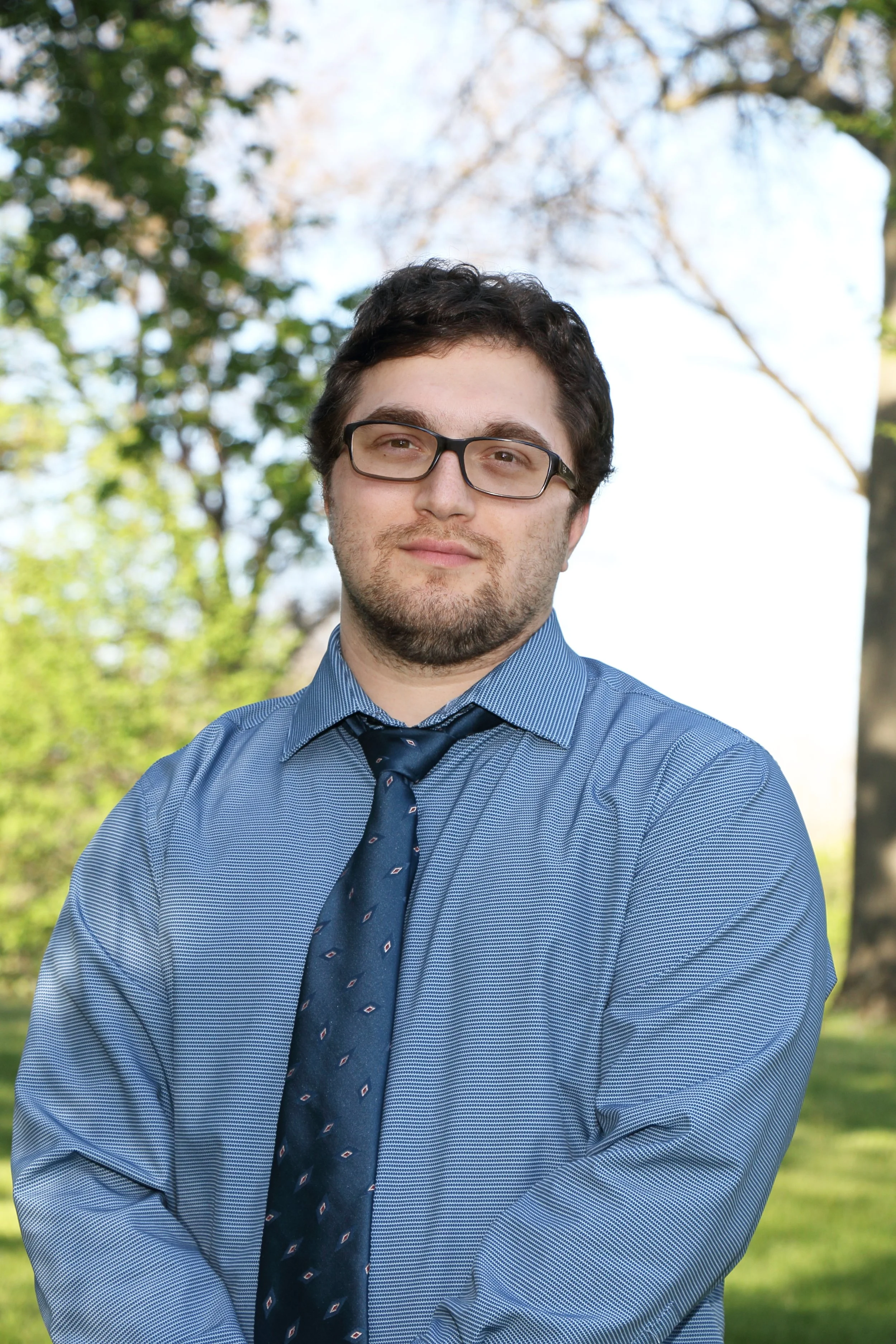 A young man with glasses and a beard in a blue dress shirt and tie, standing outdoors with trees and blue sky in the background.