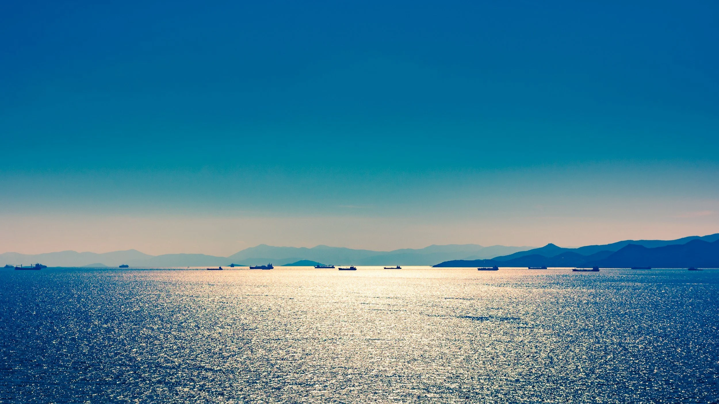 View of a vast sea with several ships sailing, distant mountain range on the horizon, and a bright blue sky.