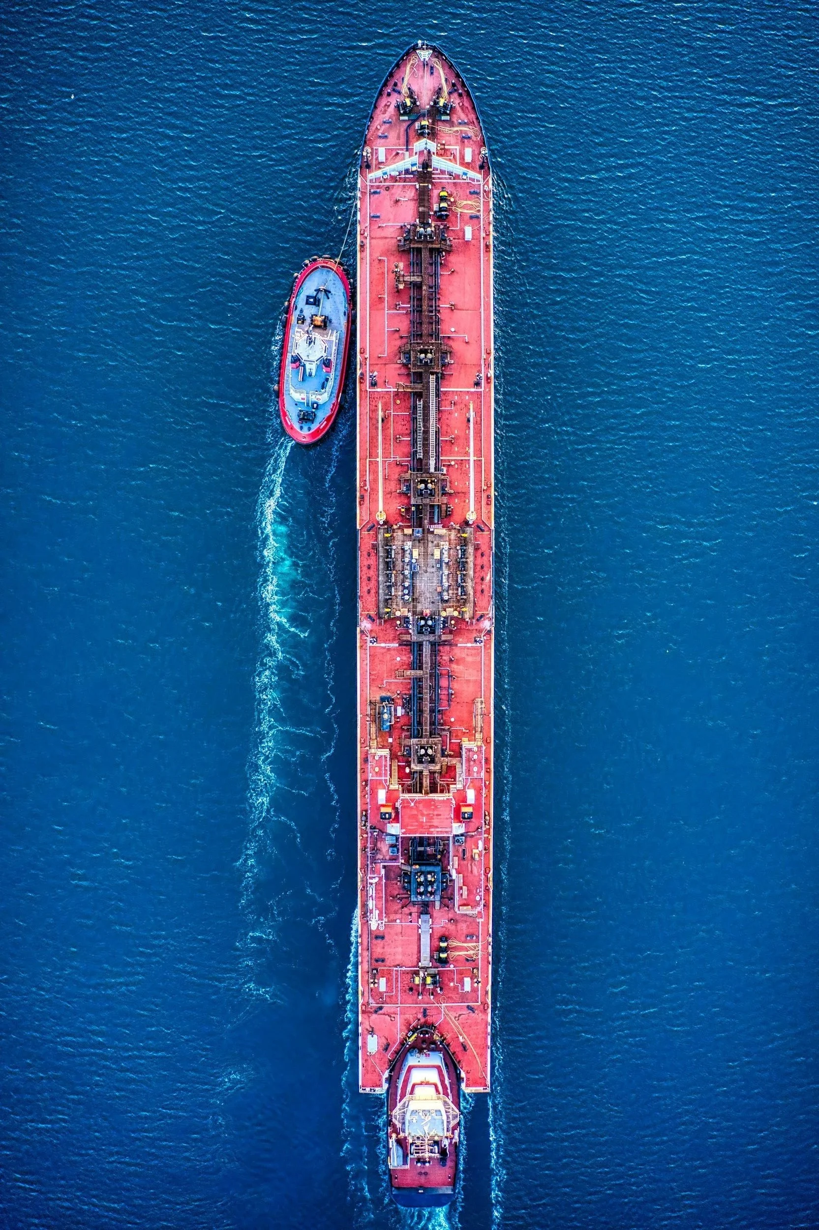 An aerial view of a large red oil tanker ship with a smaller blue and red tugboat sailing alongside in the water.