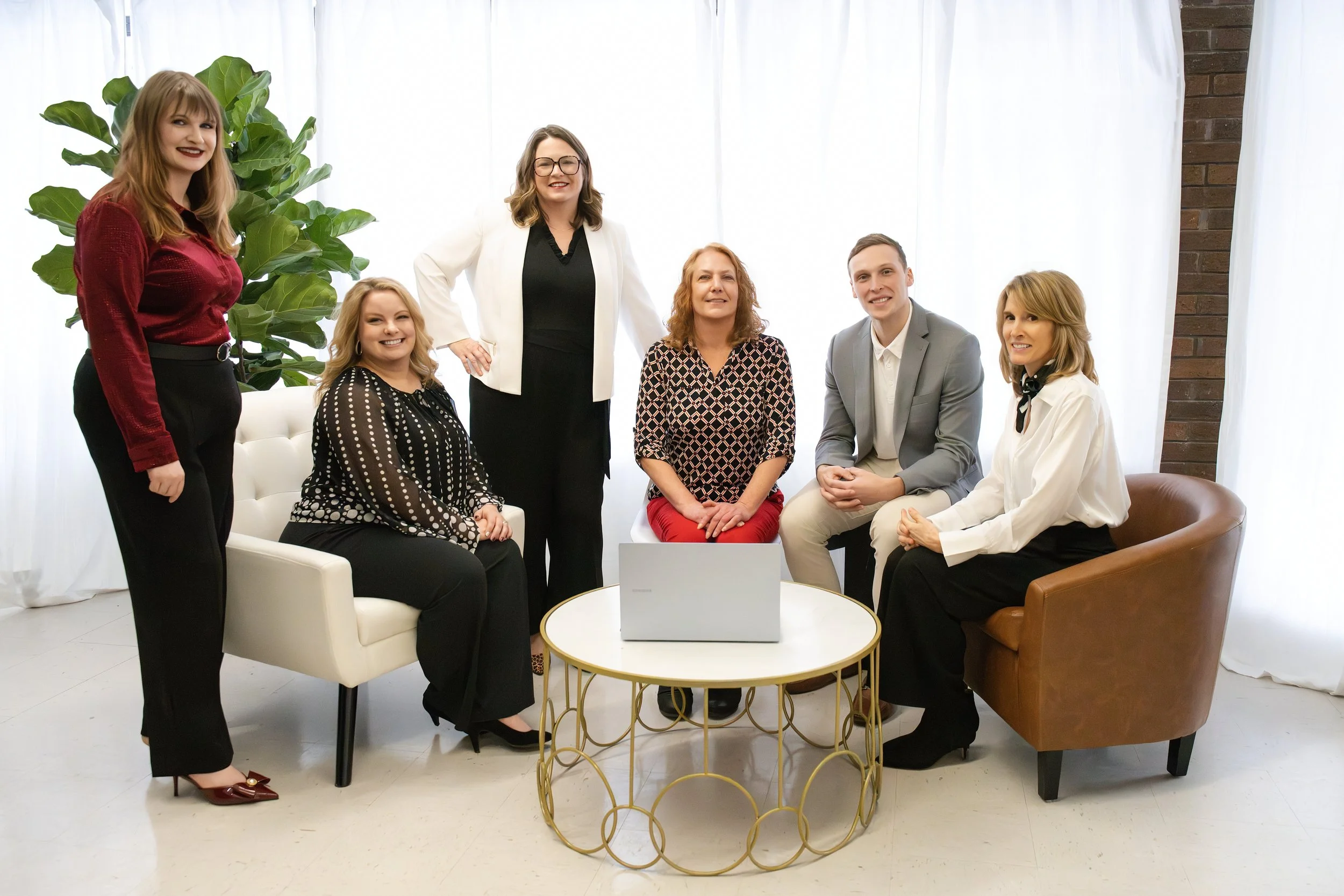 Group of seven professionals in a bright office, with white curtains and a large green plant, sitting and standing around a small round table with a laptop.