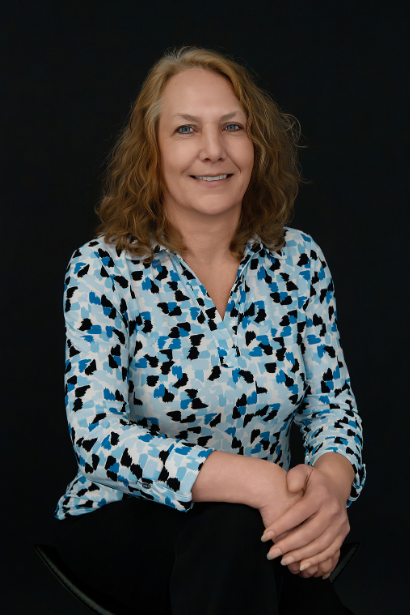 A woman with shoulder-length curly blonde hair, wearing a long-sleeved blouse with a blue, black, and white abstract pattern, sitting against a dark background.