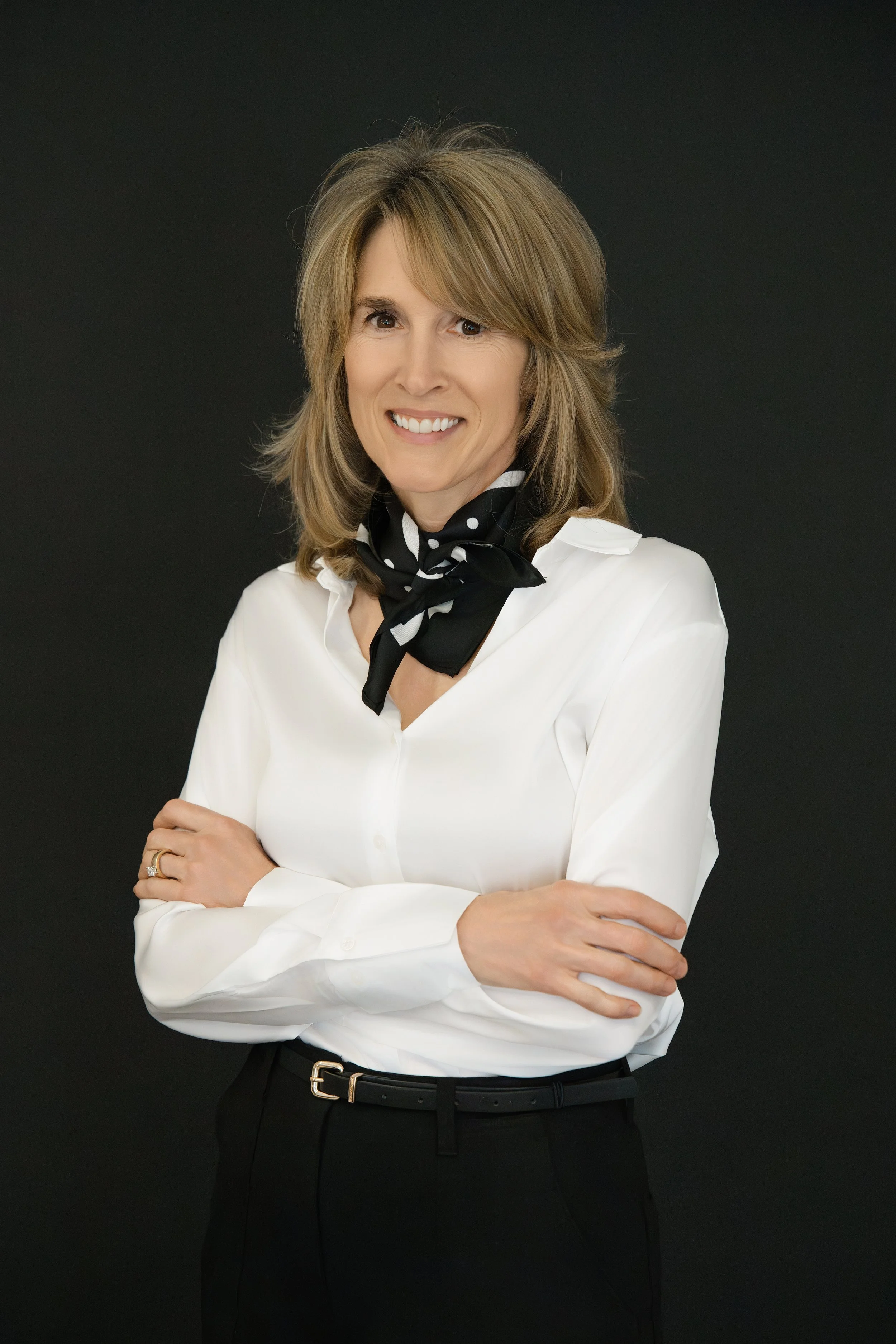 A woman with shoulder-length light brown hair, white blouse, black belt, black pants, and black polka-dotted scarf smiling against a black background.