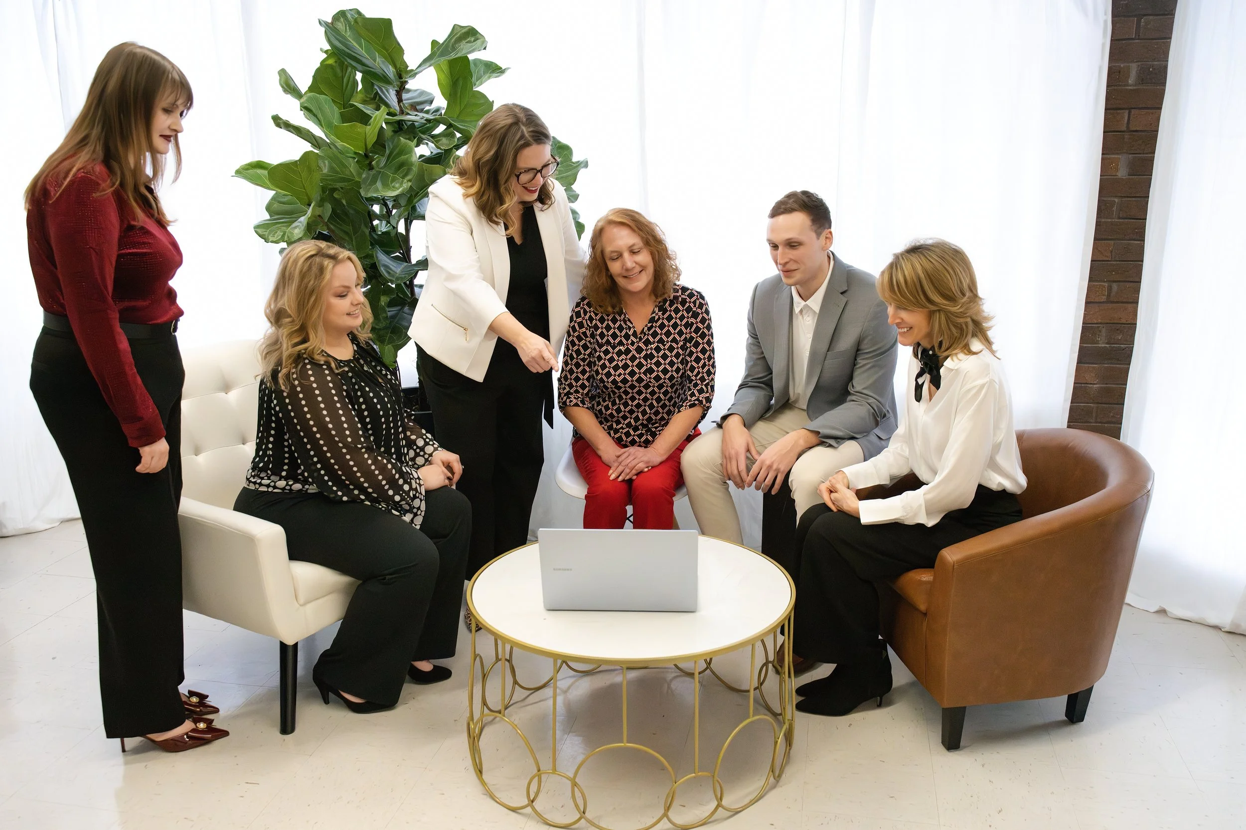 Six people gathered around a small white and gold table with a laptop, sitting and standing in a bright room with white curtains and a large green plant, engaged in a discussion or meeting.