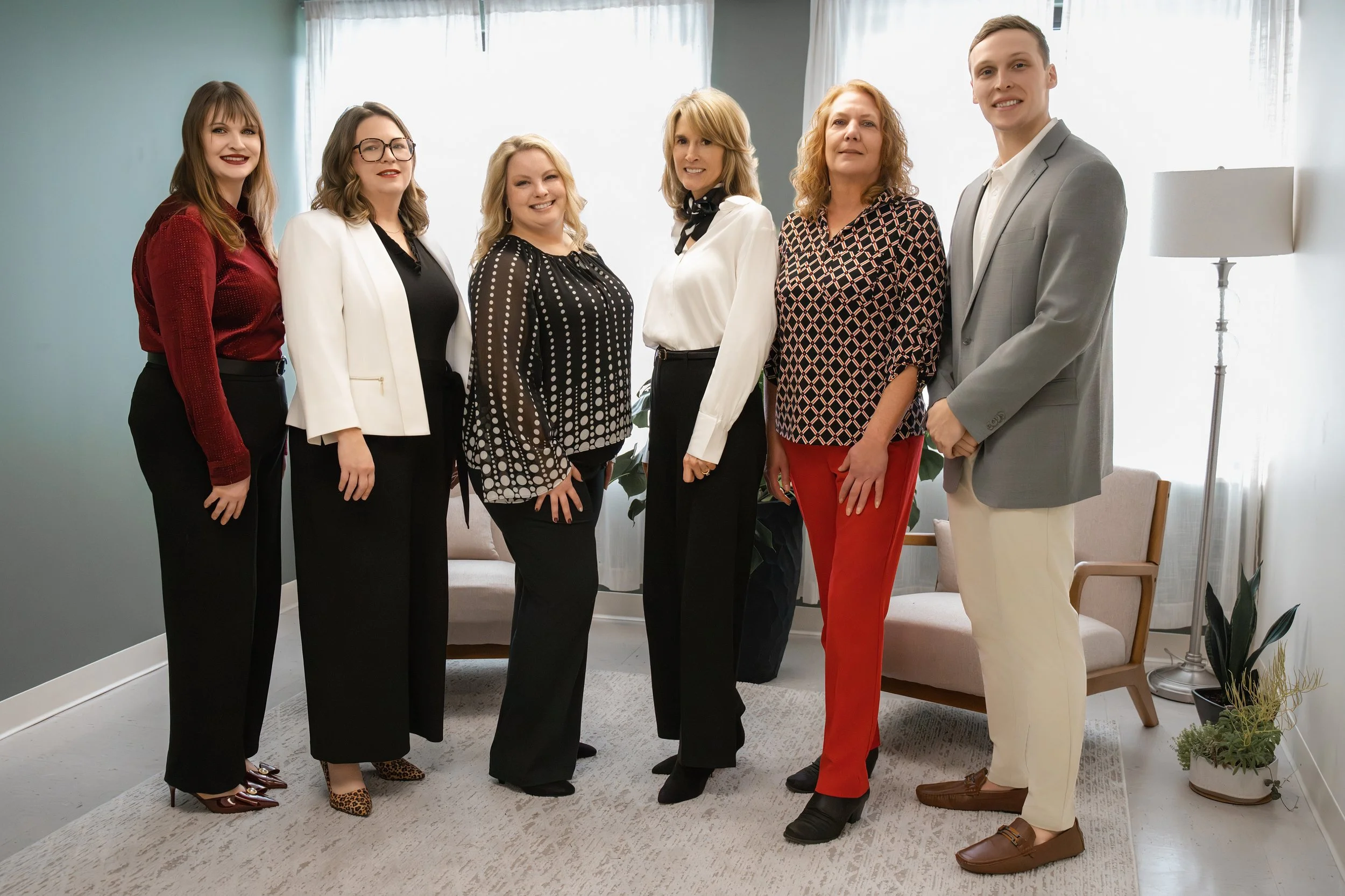 Group of seven professionally dressed women and one man standing in a living room with natural light, plants, and modern decor.