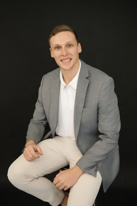 Young man wearing a gray blazer and white pants sitting against a black backdrop, smiling at the camera