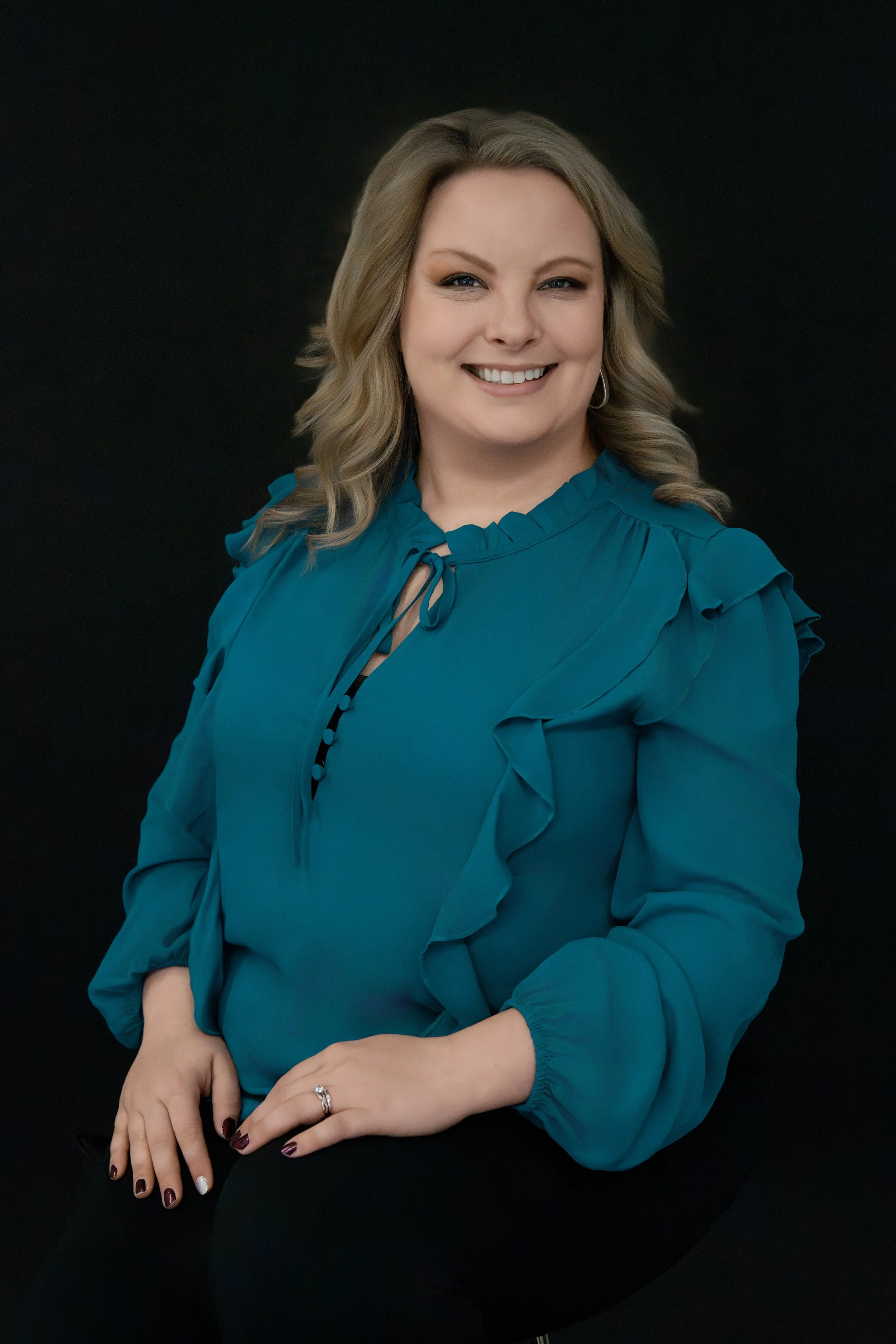 Portrait of a woman with blonde hair, smiling, wearing a teal blouse with ruffles and a bow tie, seated against a black background.
