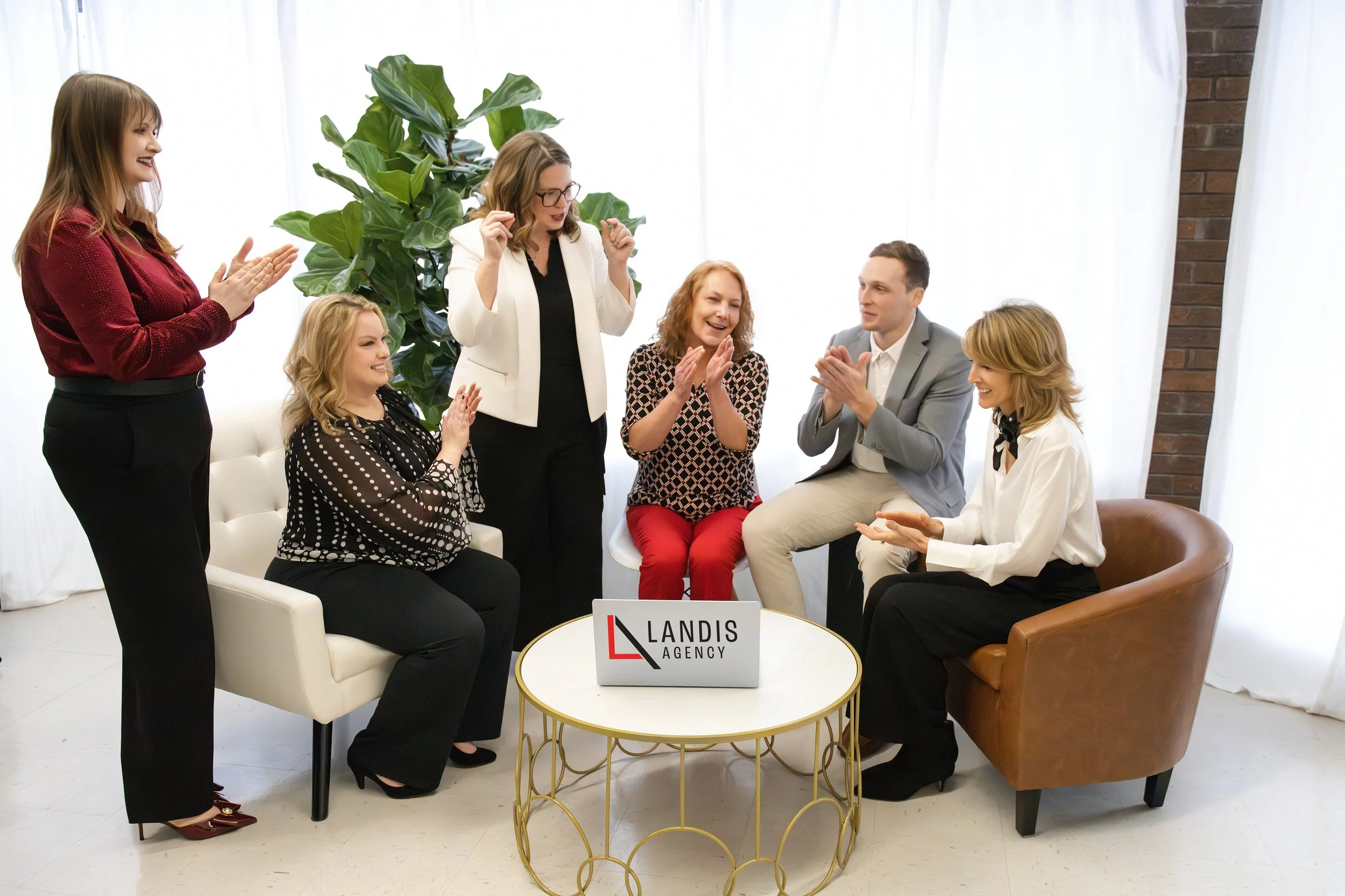 Group of six people in an office, sitting and standing around a coffee table with a laptop that reads 'Landis Agency', celebrating or having a meeting, with a large green plant in the background and white curtains.