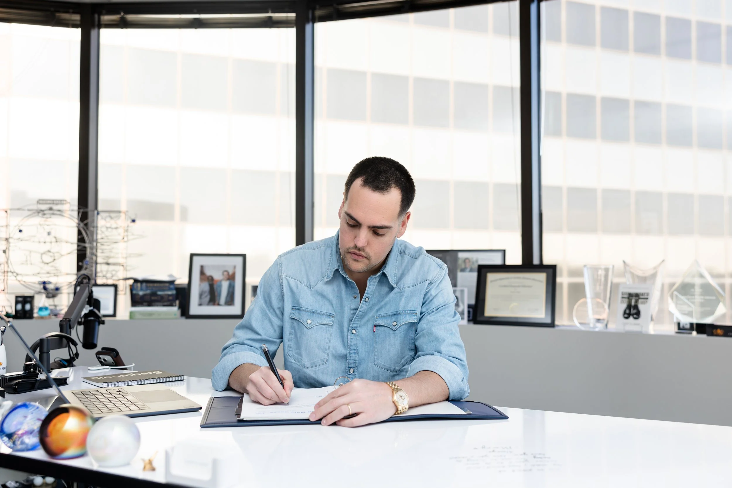 A man writing in a notebook at a white desk in an office with large windows and framed photos and certificates on the windowsill.