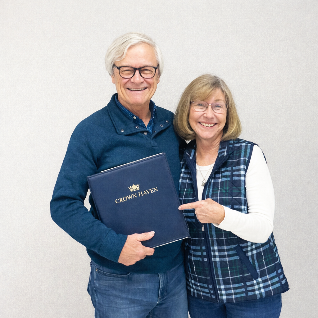 An elderly man and woman smiling, standing together, with the woman pointing at the man's folder labeled "Crown Haven".